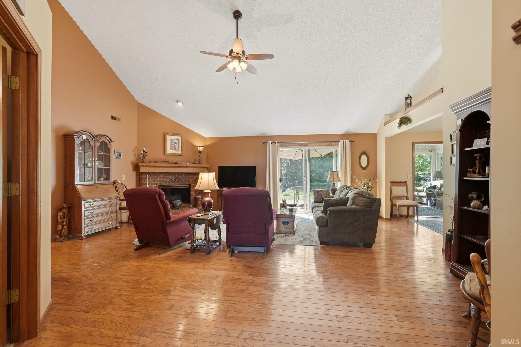 Spacious living area featuring vaulted ceilings, wood-finish flooring, a brick fireplace with a mantel, and sliding glass doors leading to the exterior