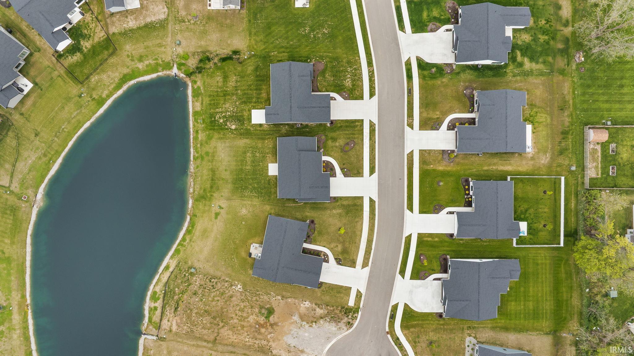 Residential community featuring a curved street, multiple single-family homes with dark-toned roofs, light-toned driveways, and green lawns, with a prominent pond bordering the property