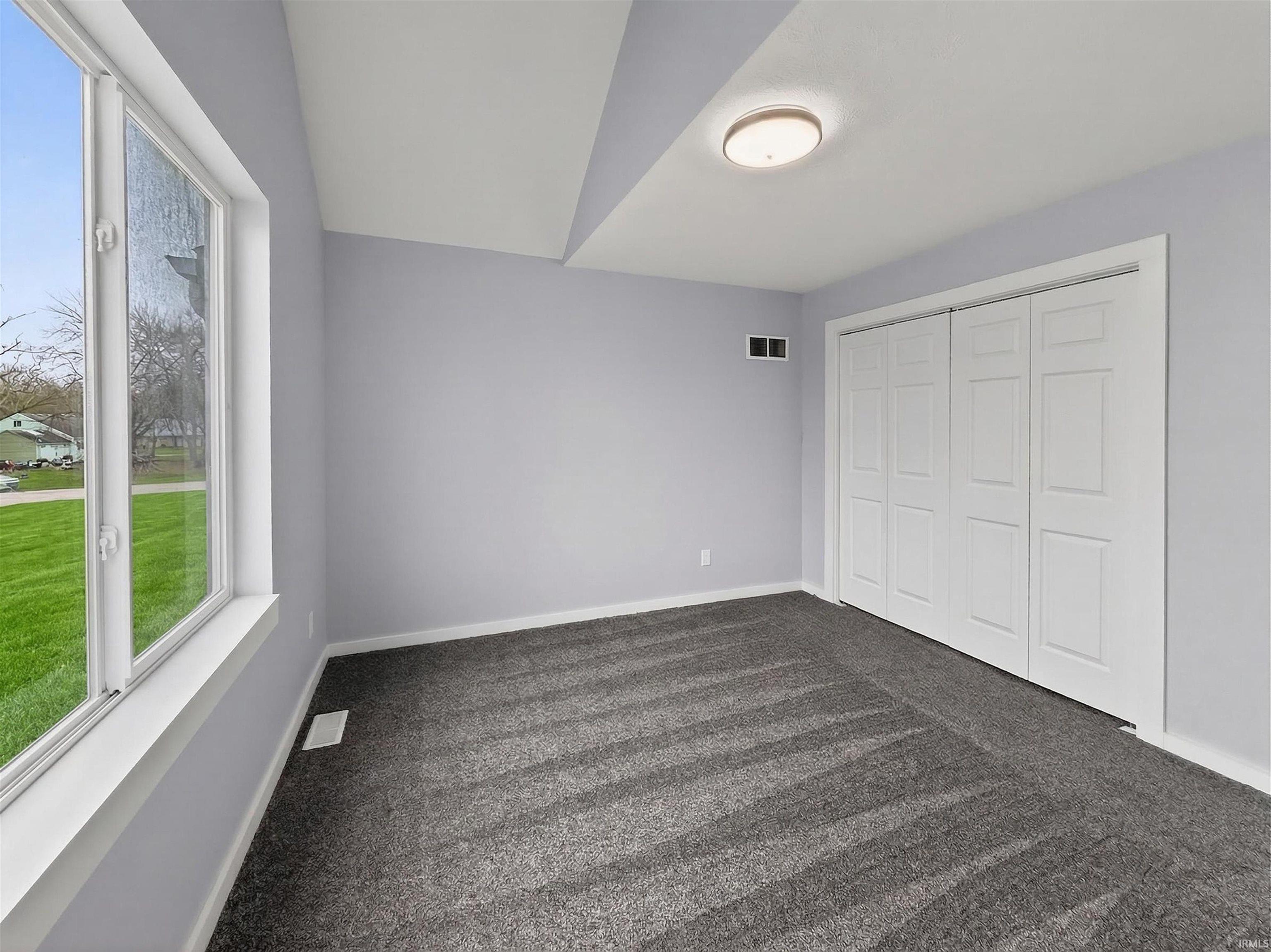 Spacious interior featuring a large window, light gray wall finishes, a built-in closet with bi-fold doors, patterned gray carpeting, and a flush-mount ceiling light fixture