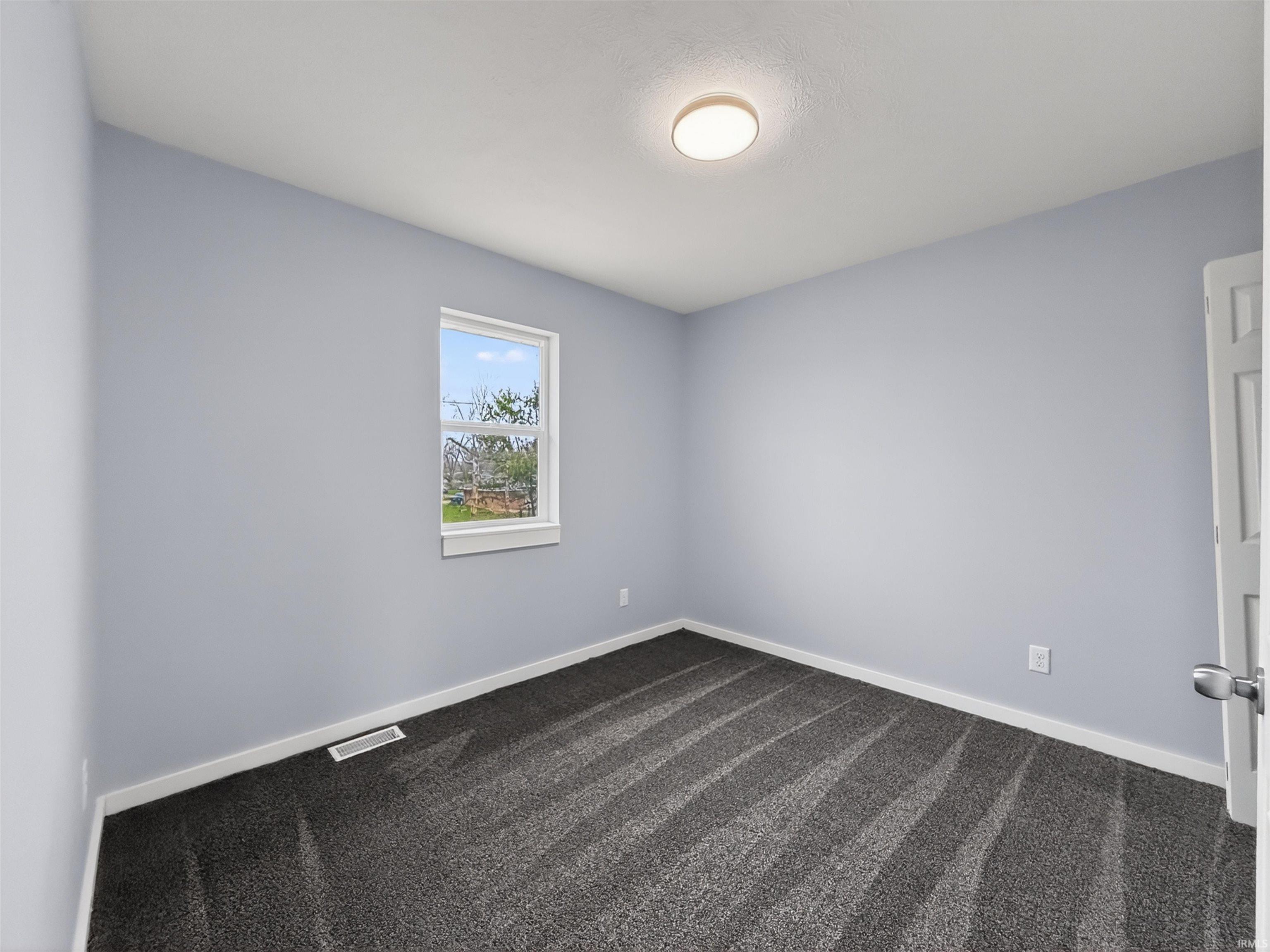Room featuring light blue walls, dark patterned carpet, a single window with white trim, a white door with a brushed nickel handle, and a flush-mount ceiling light fixture