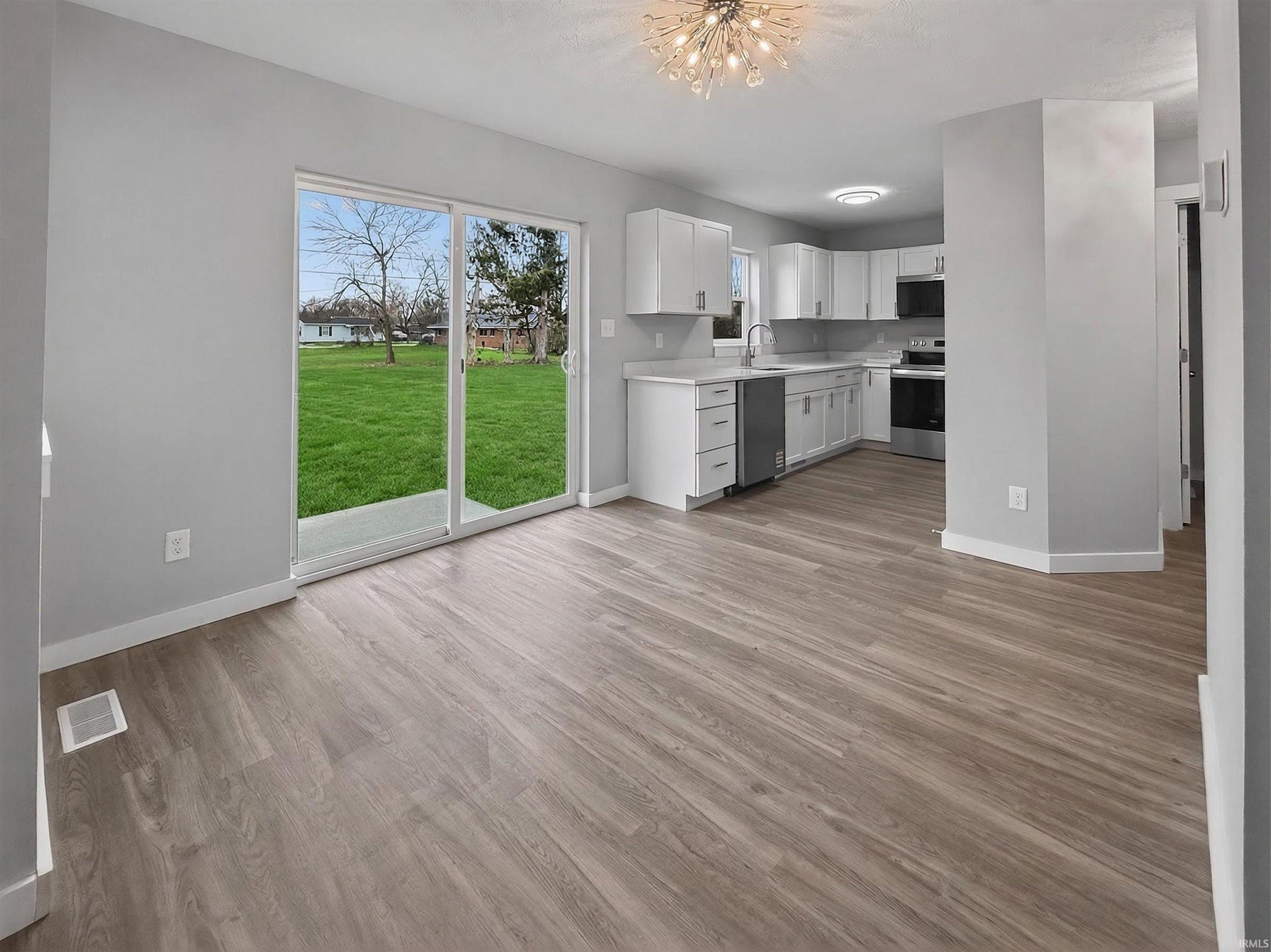 Open-concept living space featuring wood-finish flooring, a contemporary kitchen with white cabinetry and stainless steel appliances, and sliding glass doors opening to a lush green lawn