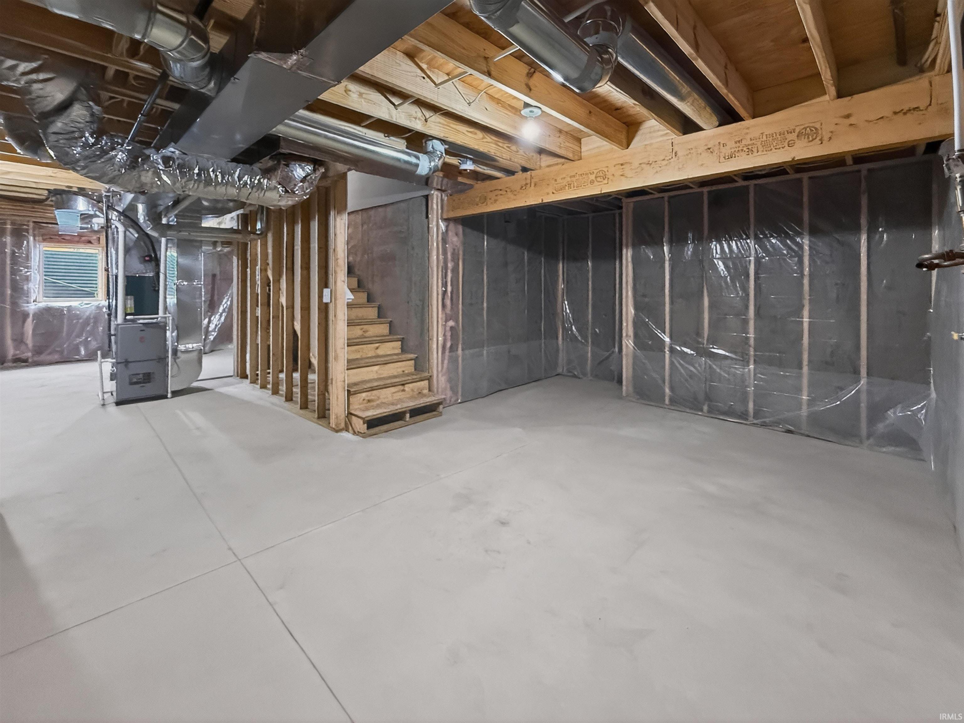 Unfinished basement featuring concrete flooring, exposed wood joists, framed walls with vapor barrier, exposed ductwork, and a built-in staircase