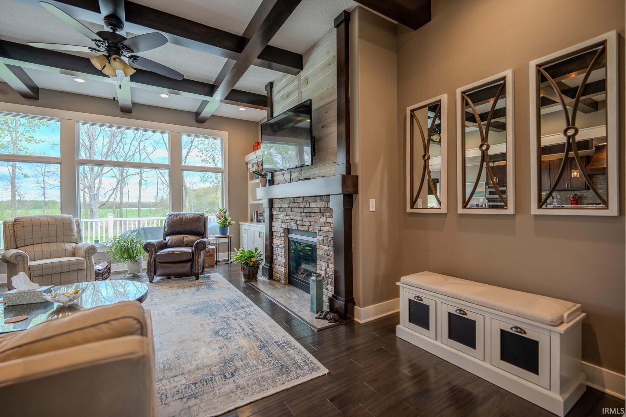 Spacious living area featuring exposed ceiling beams, a stone-faced fireplace with a wood mantle, and wood-finish flooring