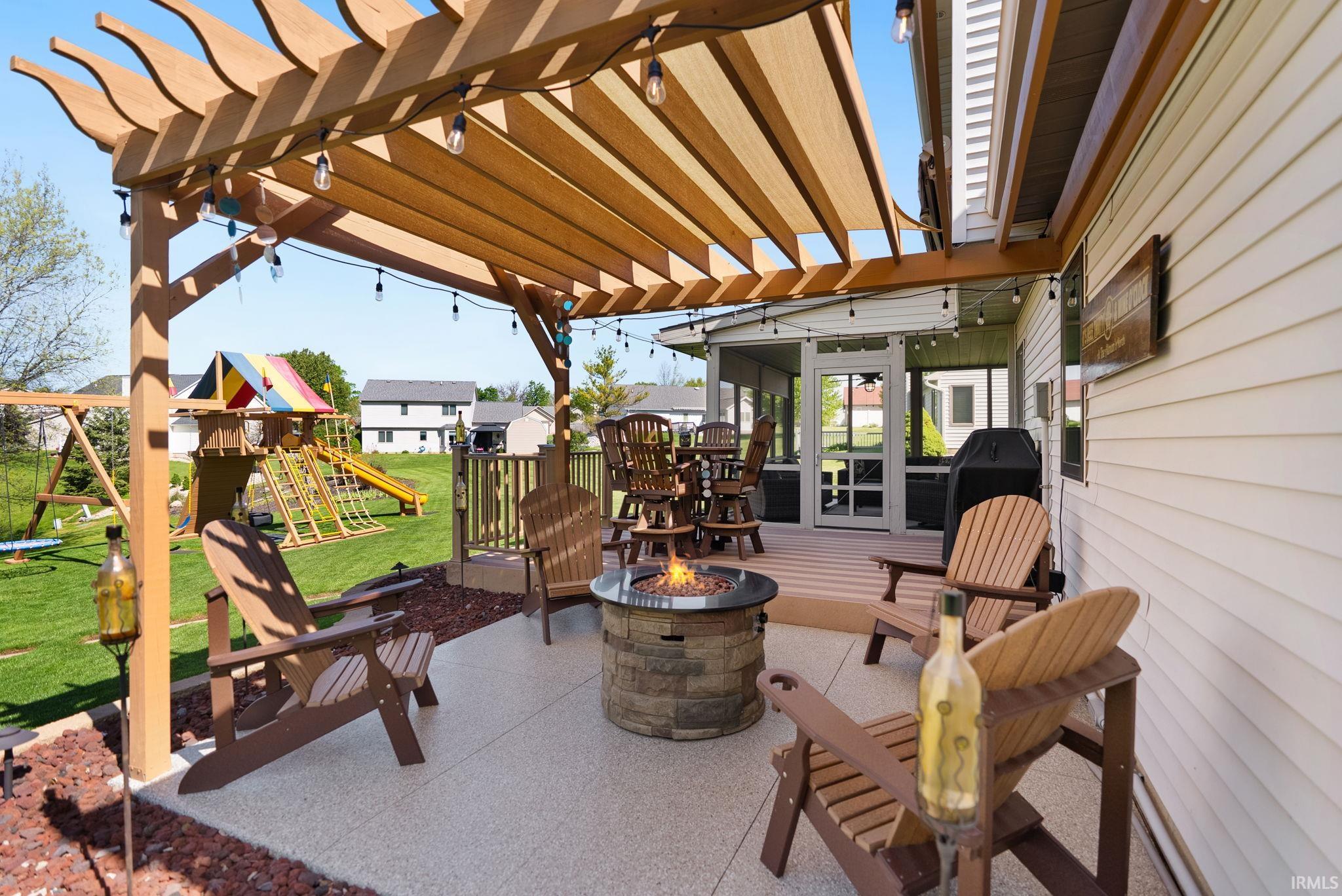 Expansive concrete patio featuring a pergola with a retractable fabric canopy, strung outdoor lighting, a stone-clad fire pit, and direct access to a screened porch with a wood-finish deck