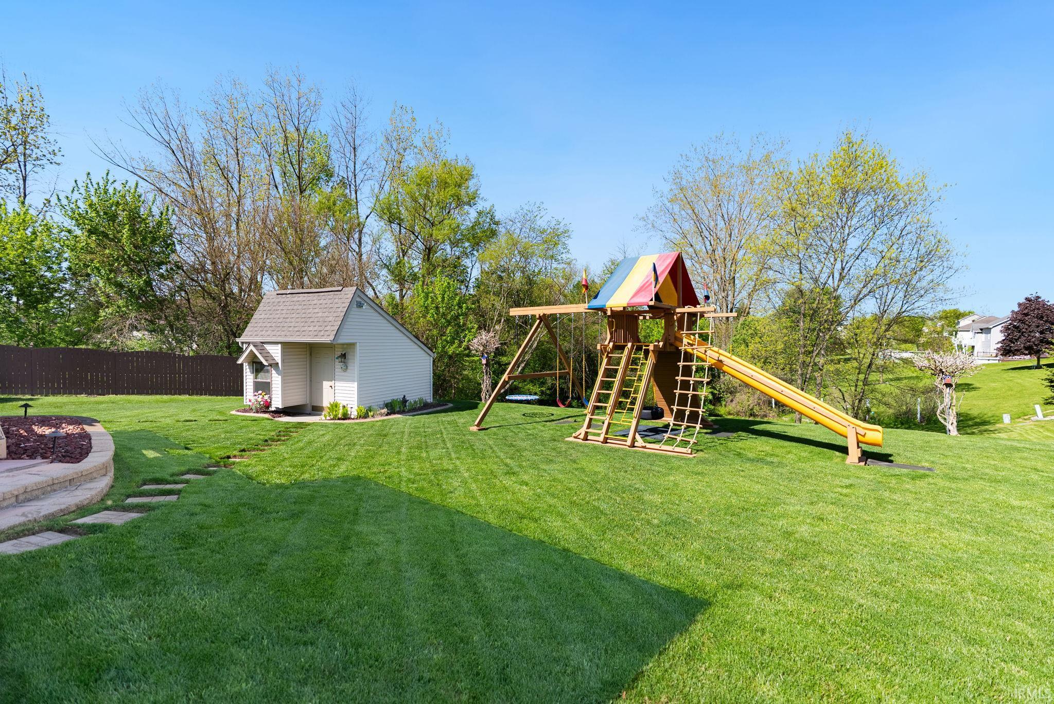 Expansive green lawn featuring a built-in fire pit with stone retaining wall, a white storage shed with shingle roof, a wooden playset with slide and climbing features, a dark wood privacy fence, and mature trees