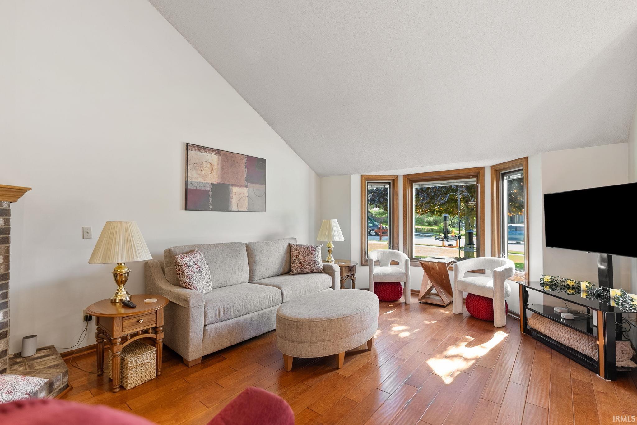 Spacious living area featuring vaulted ceilings, wood-finish flooring, a brick fireplace surround with a wood mantel, and large bay windows