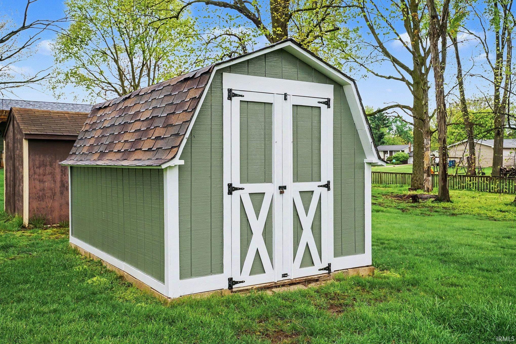 Green storage shed featuring a gambrel roof with shingle siding, white trim, and white double barn doors with black hardware