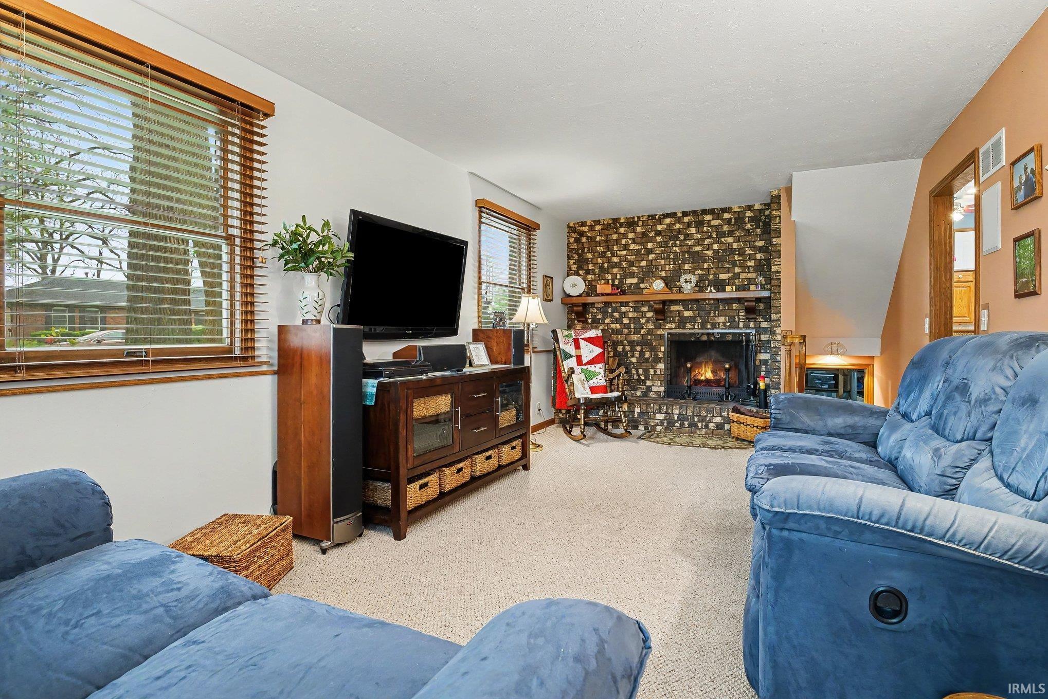 Living area featuring a brick-surround fireplace with a wood mantel, light-colored carpet flooring, and a large window with wood-finish blinds