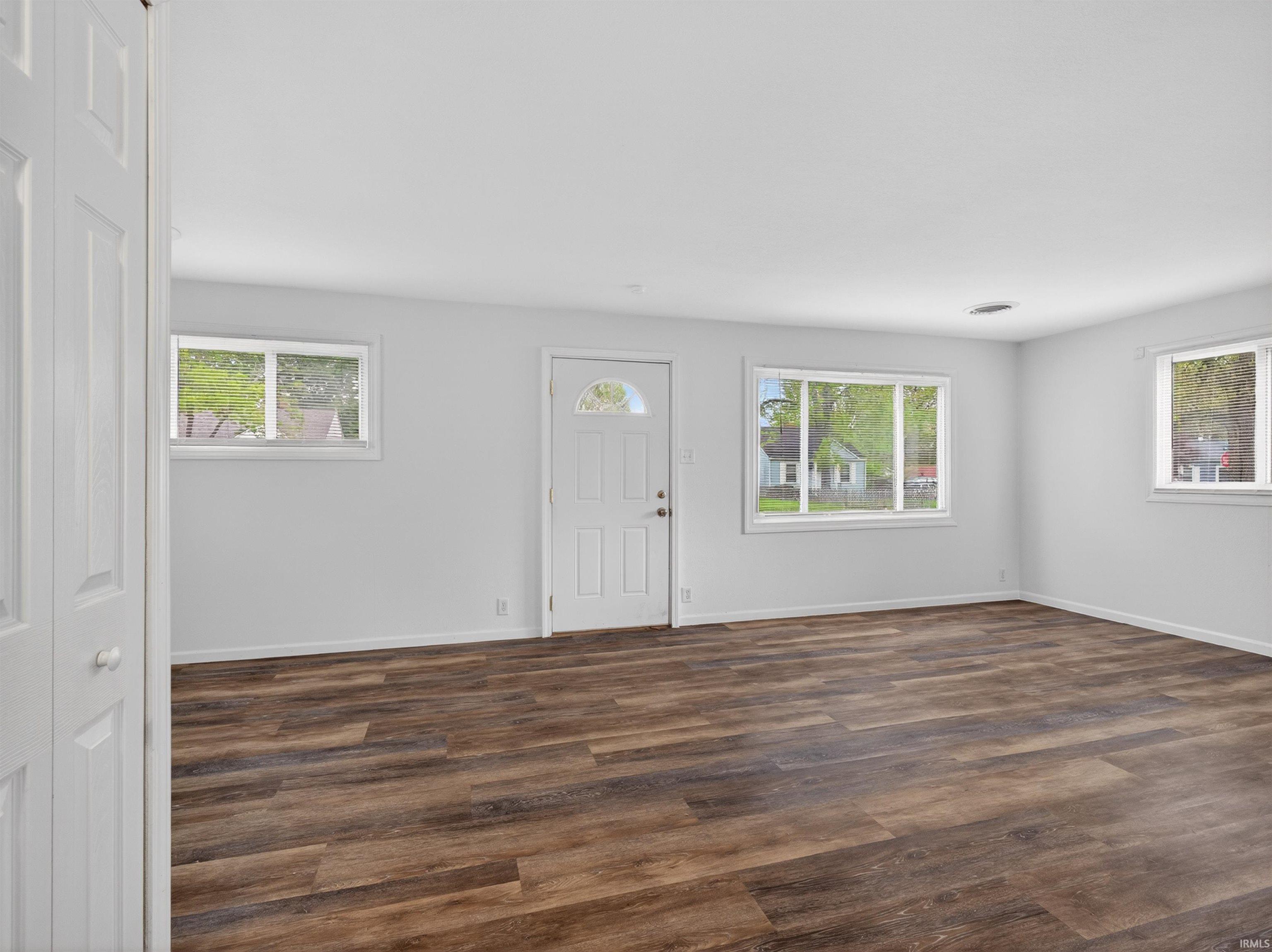 Spacious room featuring wood-finish flooring, white walls, multiple windows, and a panel door with an arched glass inset