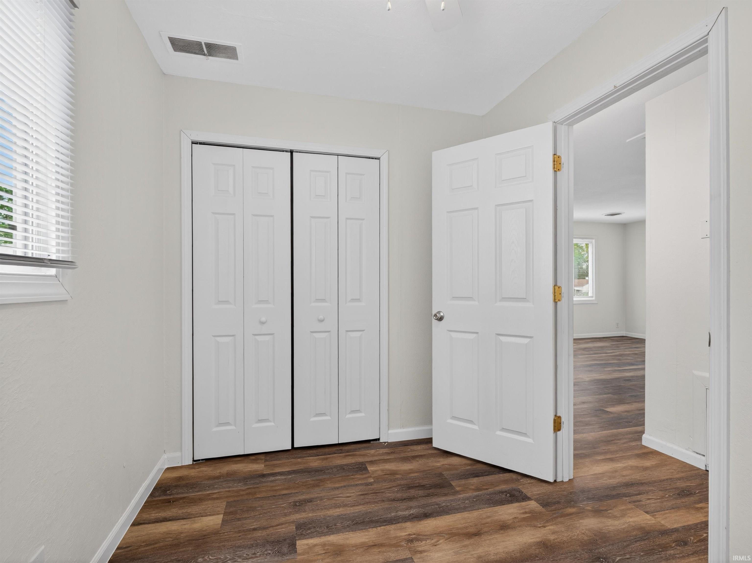 Room featuring wood-finish flooring, white bi-fold closet doors, a window with horizontal blinds, and light-colored walls