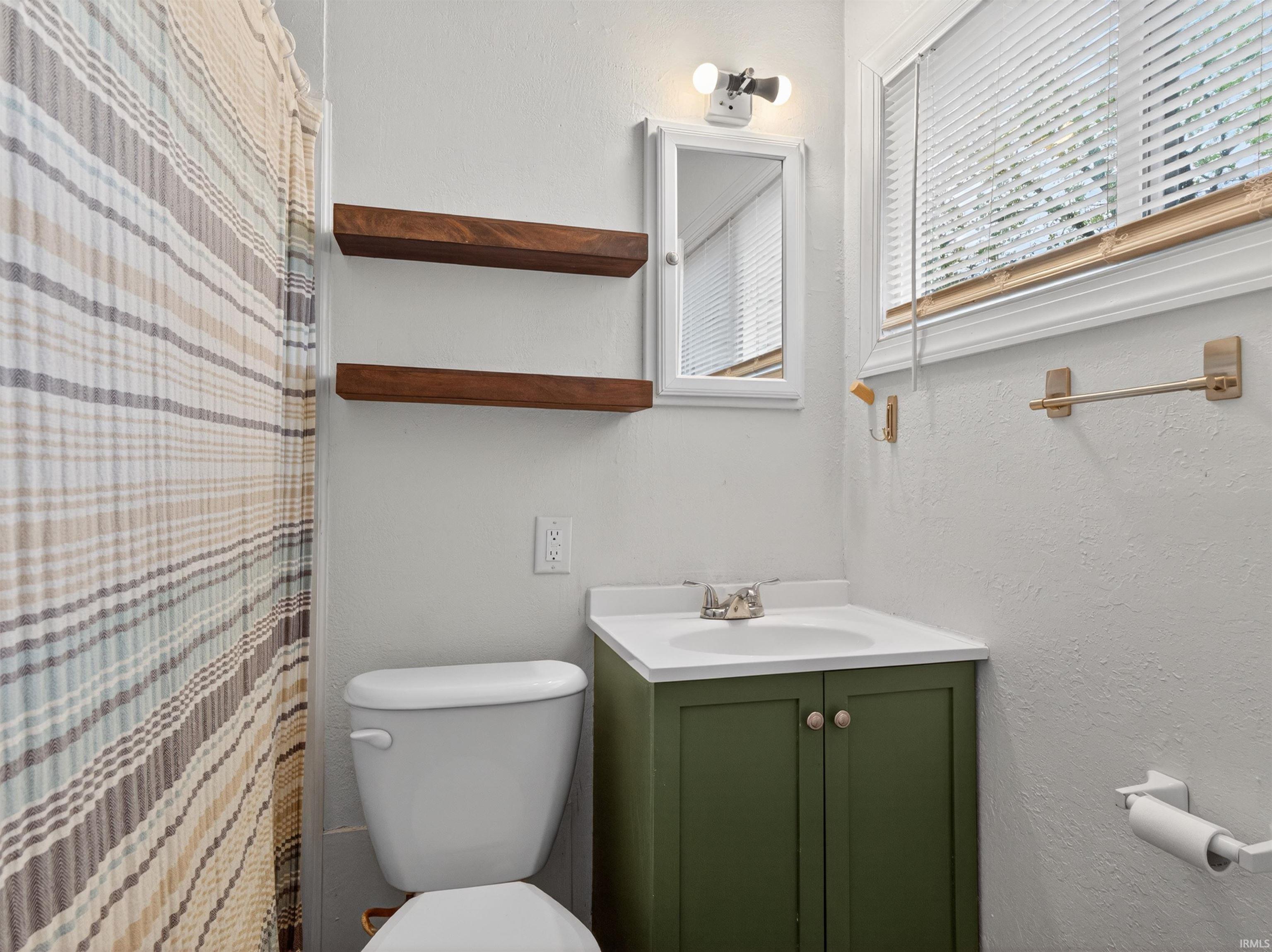 Bathroom featuring a green vanity with a white countertop, two floating wood shelves, a built-in medicine cabinet, and a window with blinds