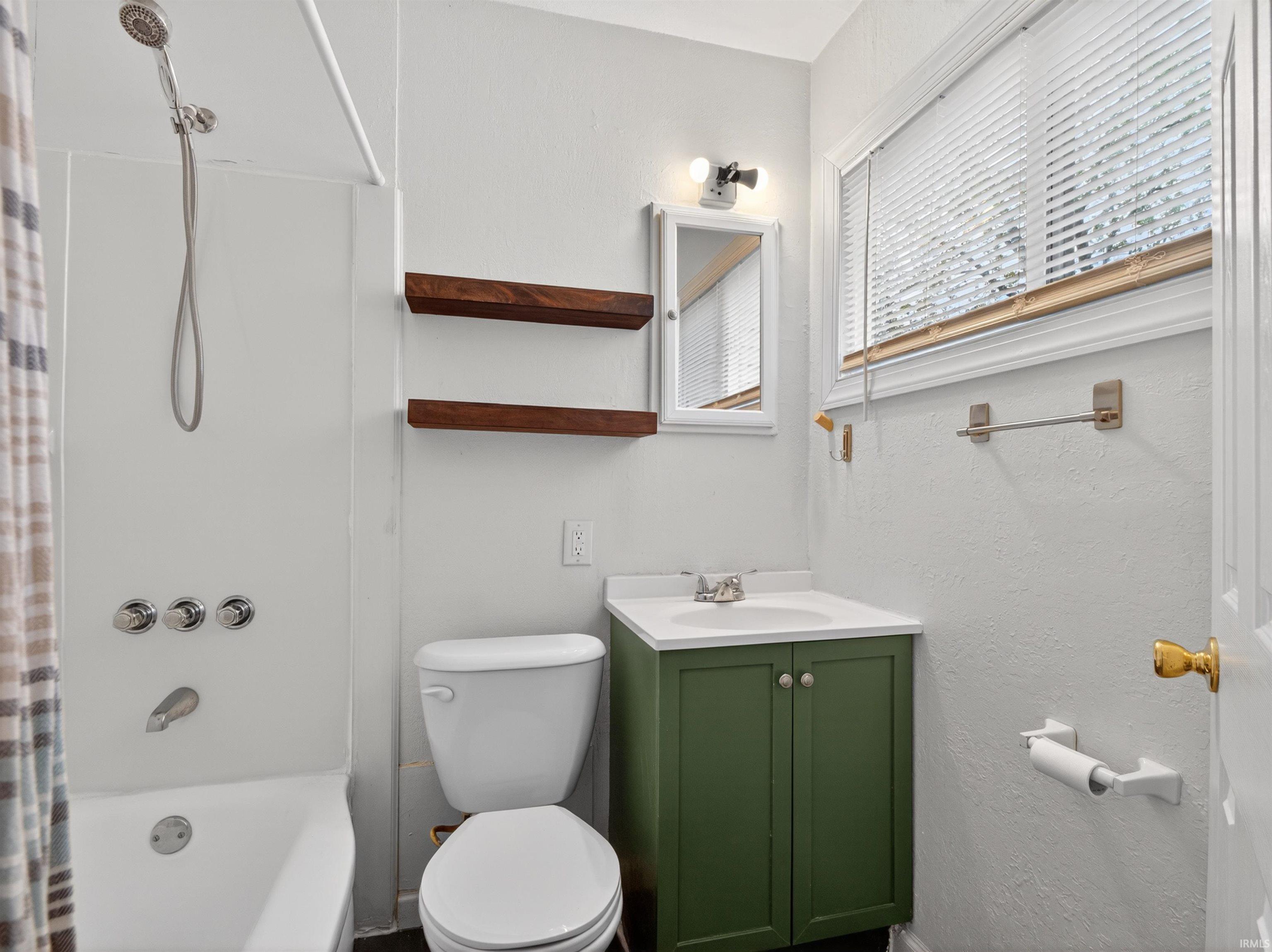 Bathroom featuring a white tub with shower head, two wood-finish floating shelves, a vanity with a green cabinet and integrated sink, and a window with blinds