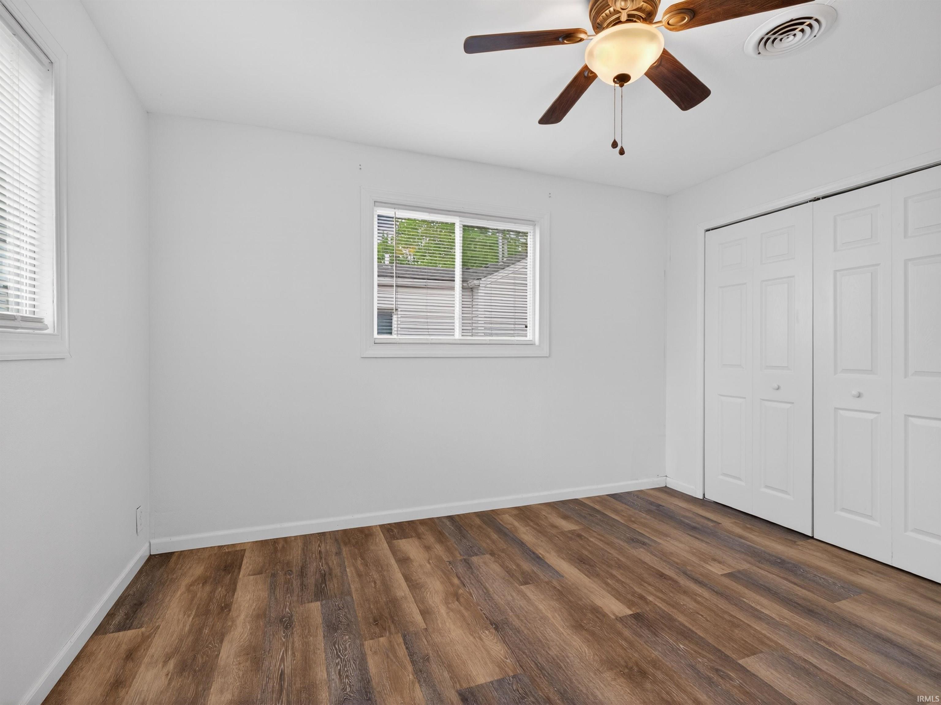 Bedroom featuring wood-finish flooring, two windows with blinds, a ceiling fan with integrated lighting, and bi-fold closet doors
