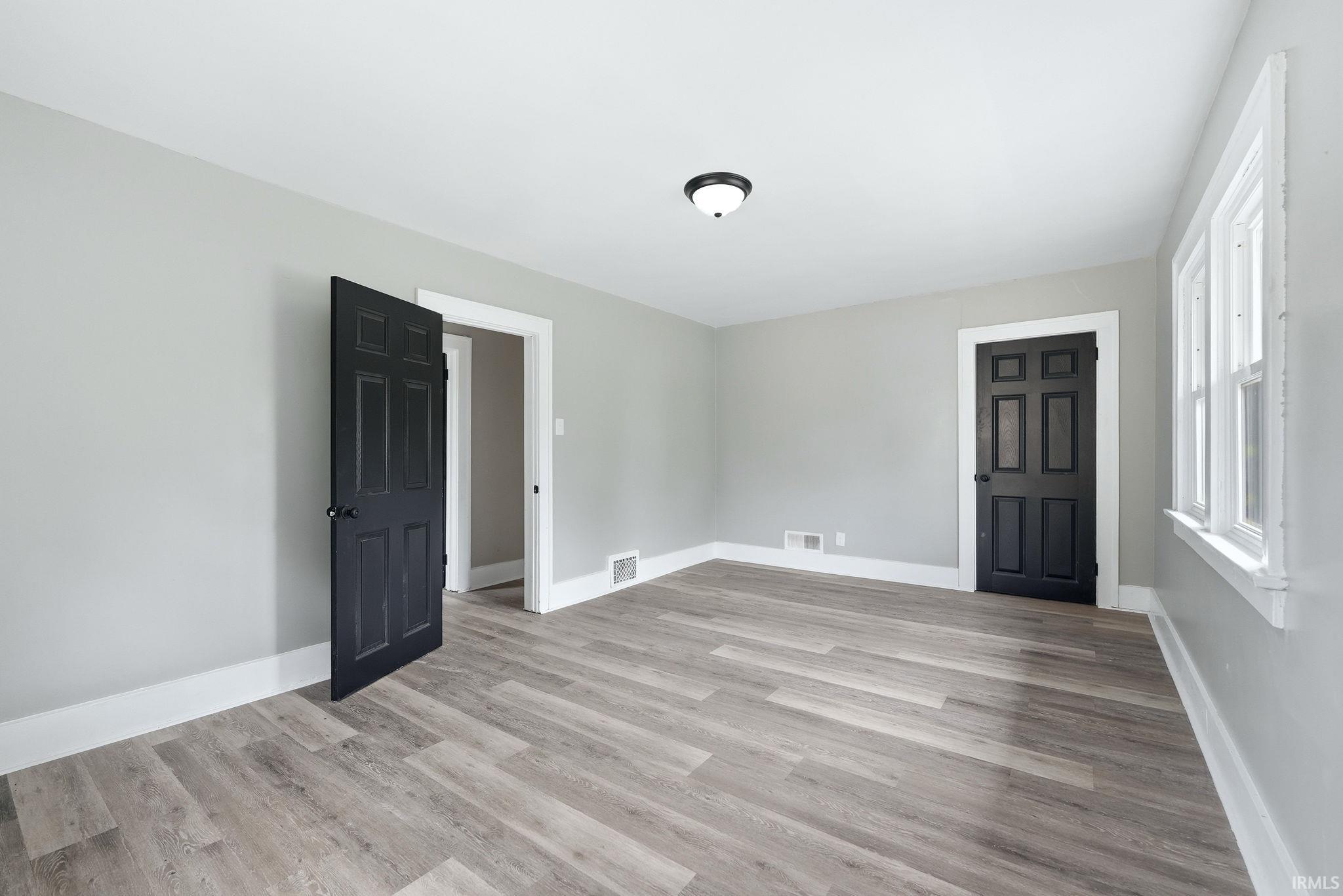 Spacious room featuring wood-finish flooring, light gray walls, white trim, a multi-pane window, and black paneled doors