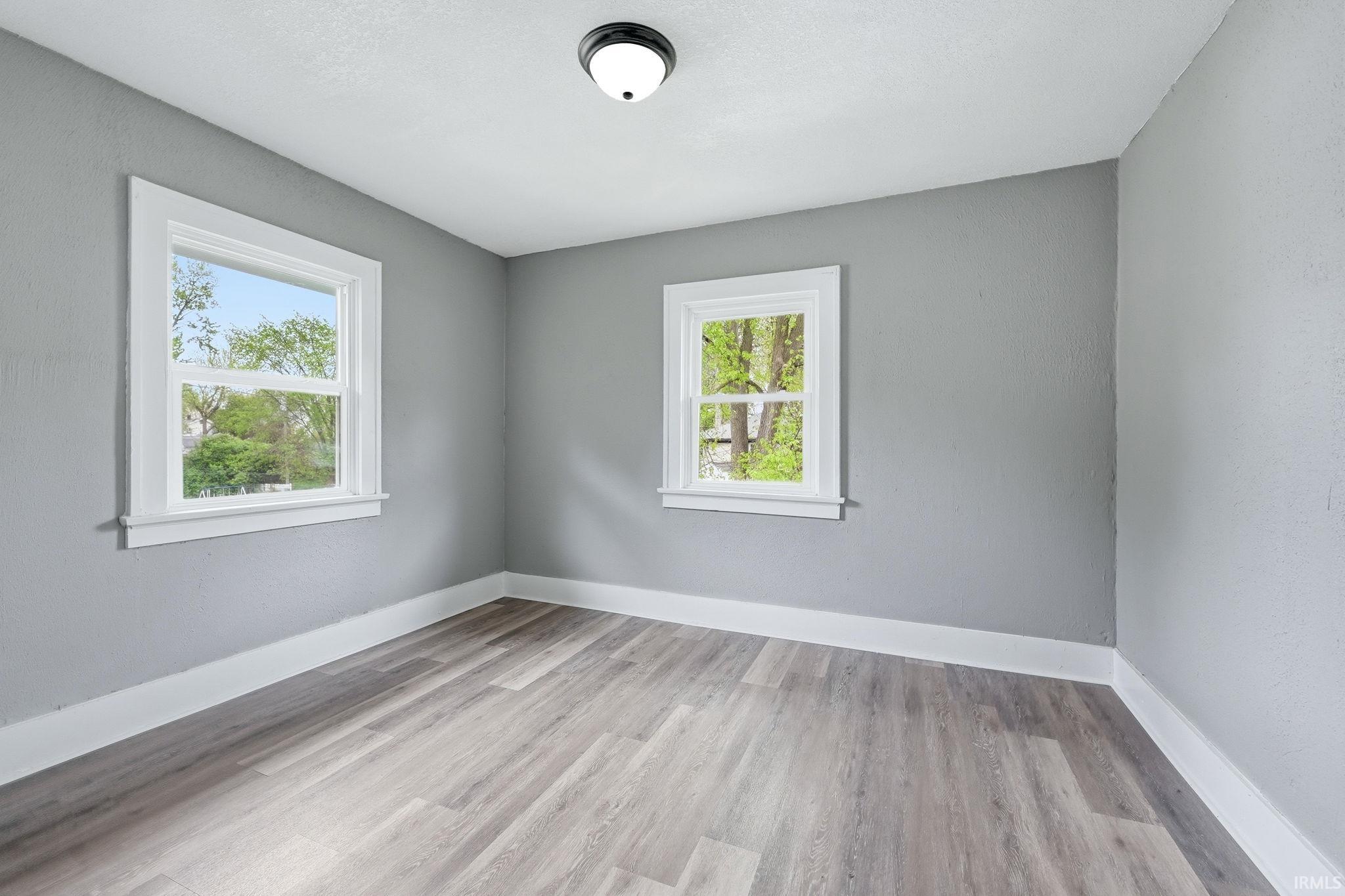 Bright room featuring two double-hung windows with white trim, light gray walls, wood-finish flooring, white baseboards, and a flush-mount ceiling light fixture