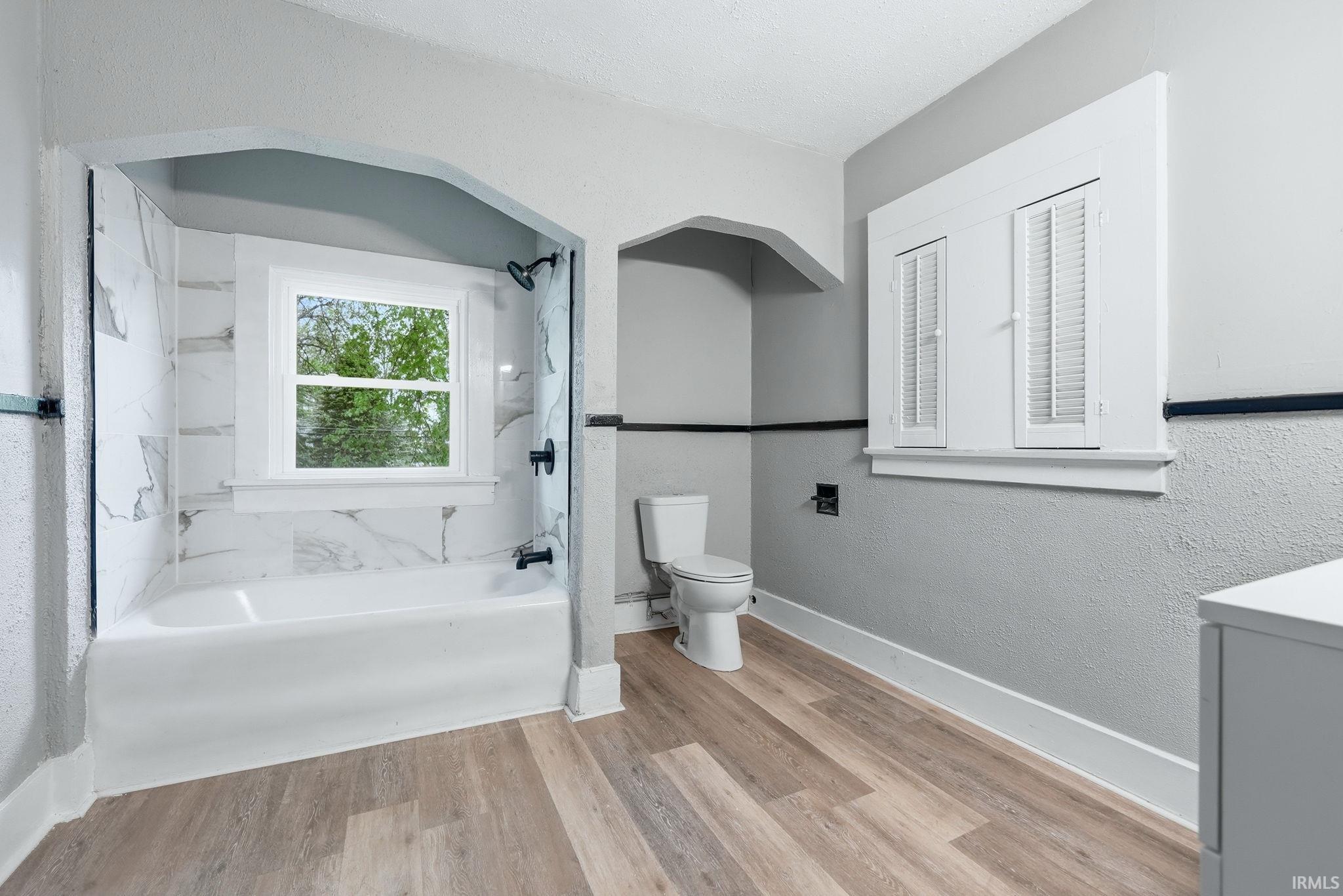 Bathroom featuring wood-finish flooring, a white bathtub with marble-look tile surround, a window with exterior views, and a toilet