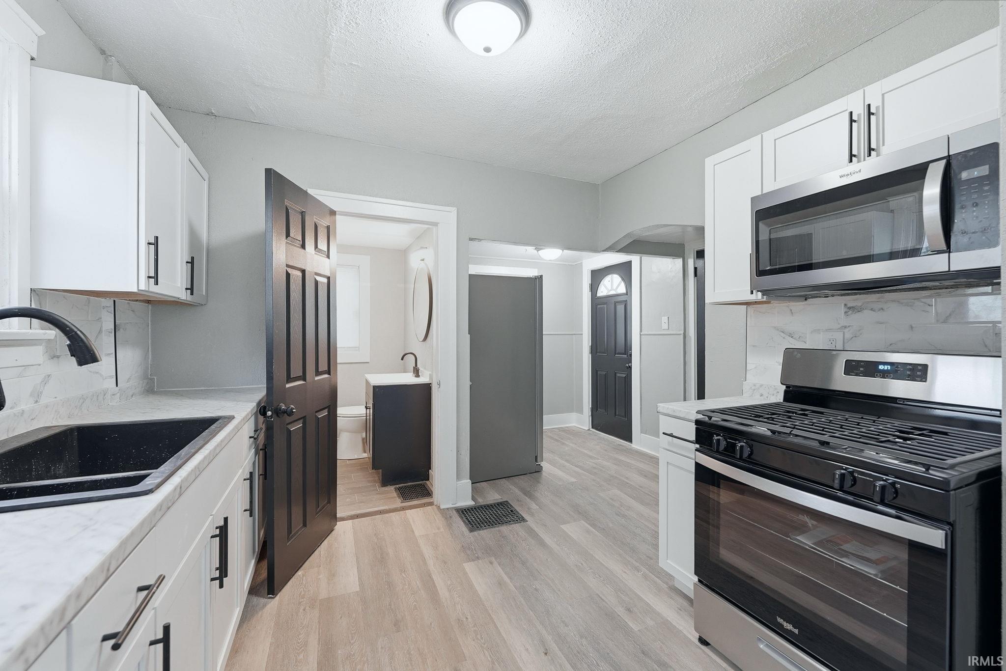 Modern kitchen featuring white cabinetry with matte black hardware, light-toned wood-finish flooring, a black composite sink with a matte black faucet, white countertops, and a stainless steel gas range with an over-the-range microwave