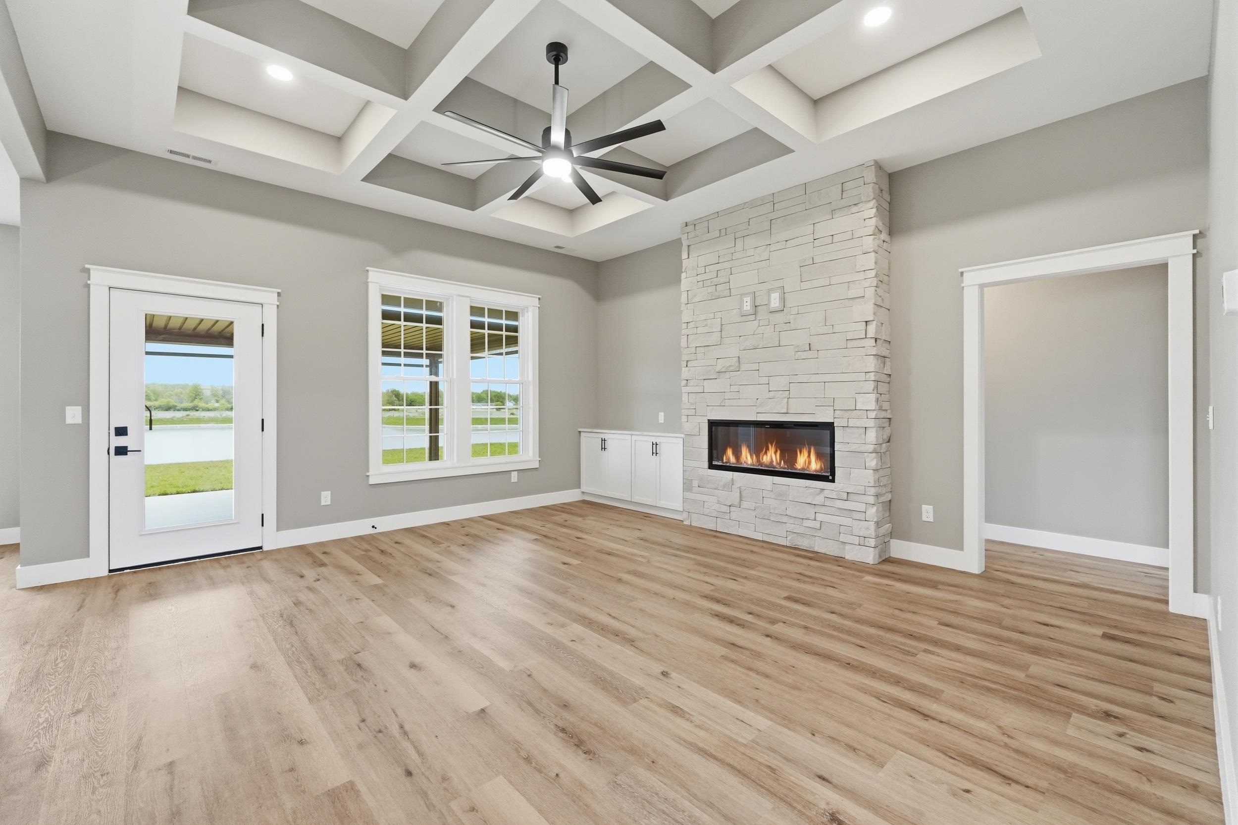 Spacious room featuring wood-finish flooring, a contemporary stacked stone fireplace, and a coffered ceiling with a ceiling fan
