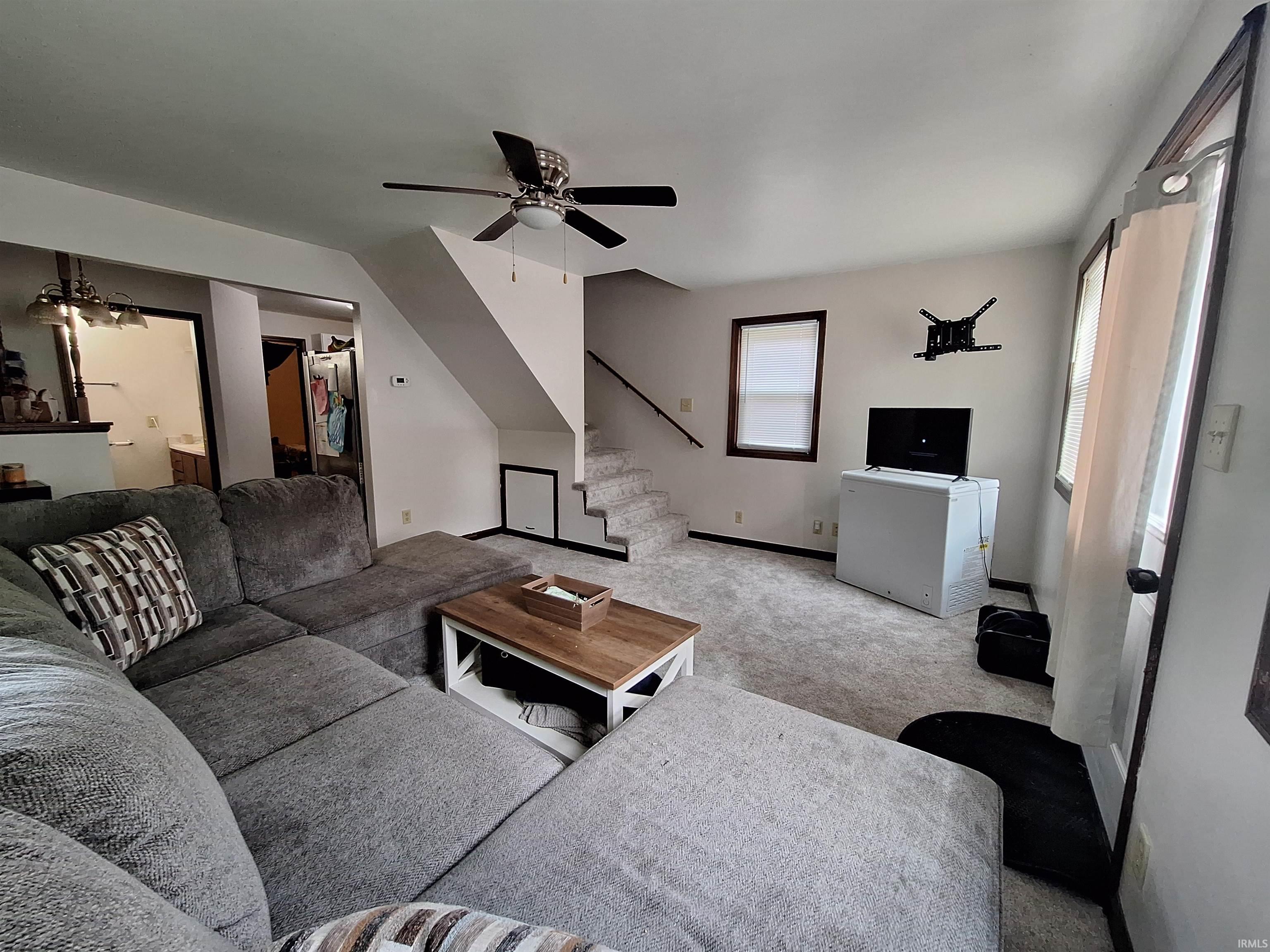 Carpeted living area featuring a ceiling fan, staircase, and natural light from multiple windows