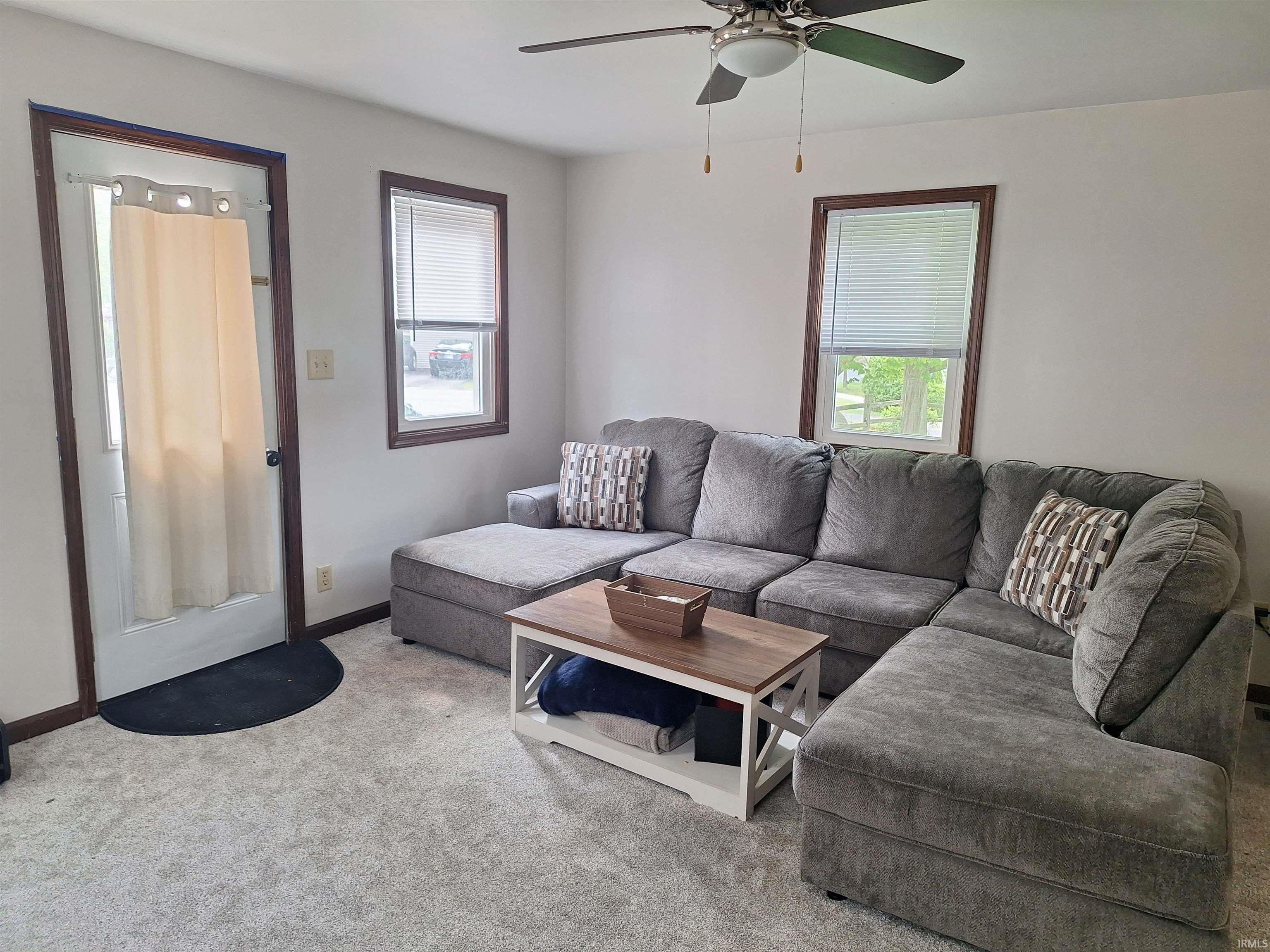 Living area featuring light-colored carpet flooring, white walls, and a ceiling fan with integrated lighting