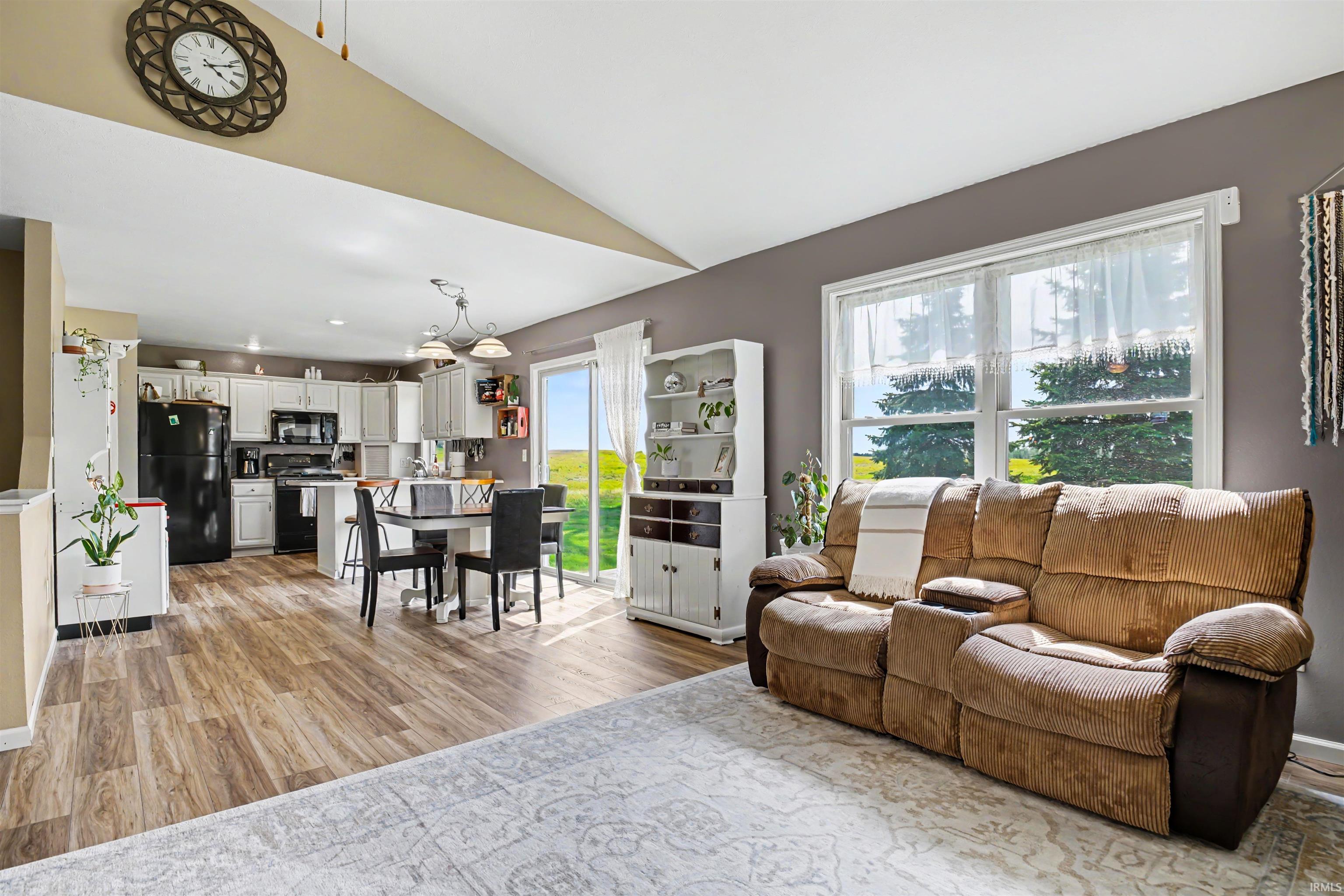 Open-concept living area featuring wood-finish flooring, a vaulted ceiling, and a kitchen with white cabinetry and black appliances