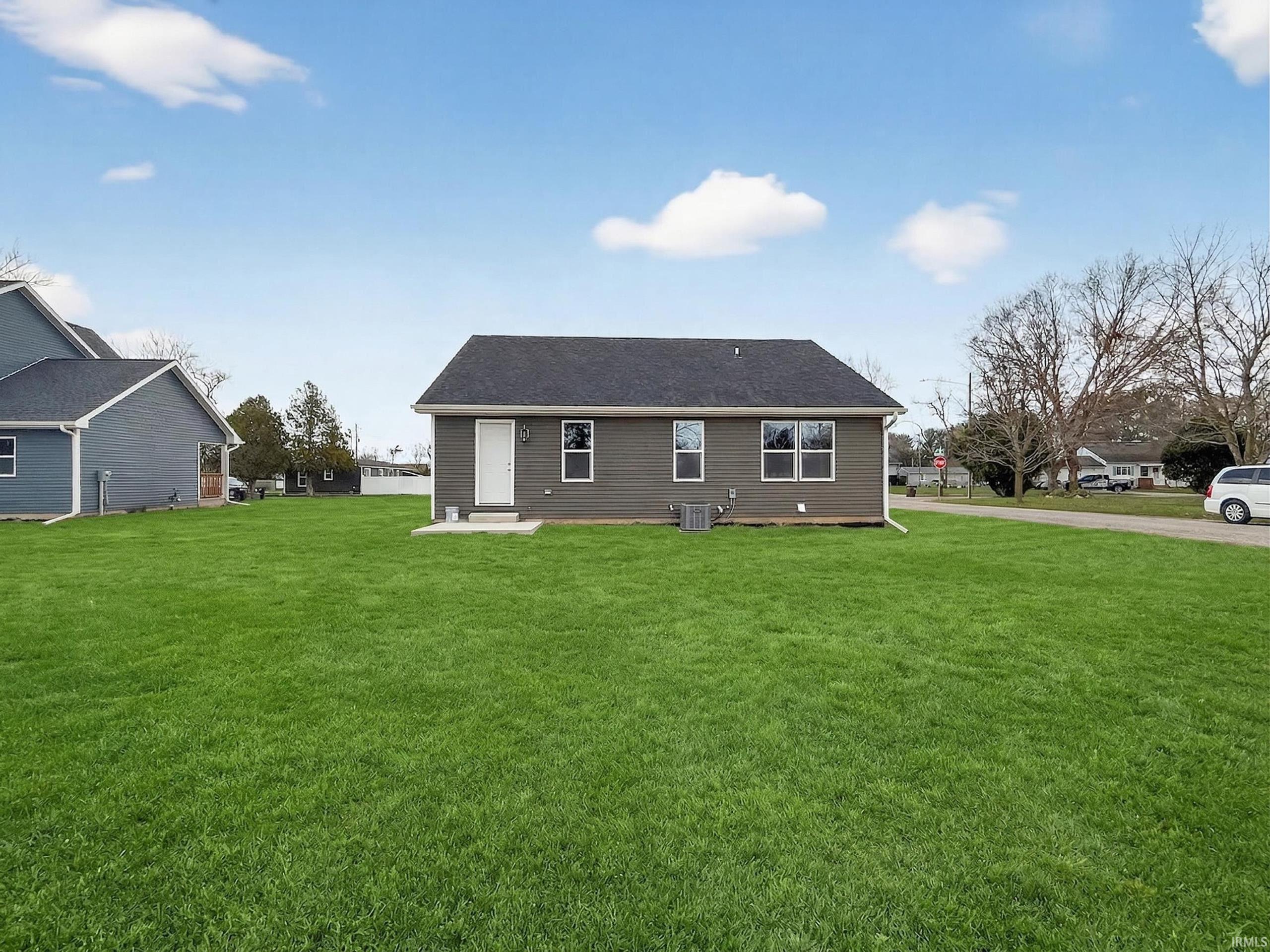 Expansive green lawn with a single-story dwelling featuring gray horizontal siding, multiple windows, a dark shingle roof, and a white rear entrance door with a small concrete pad