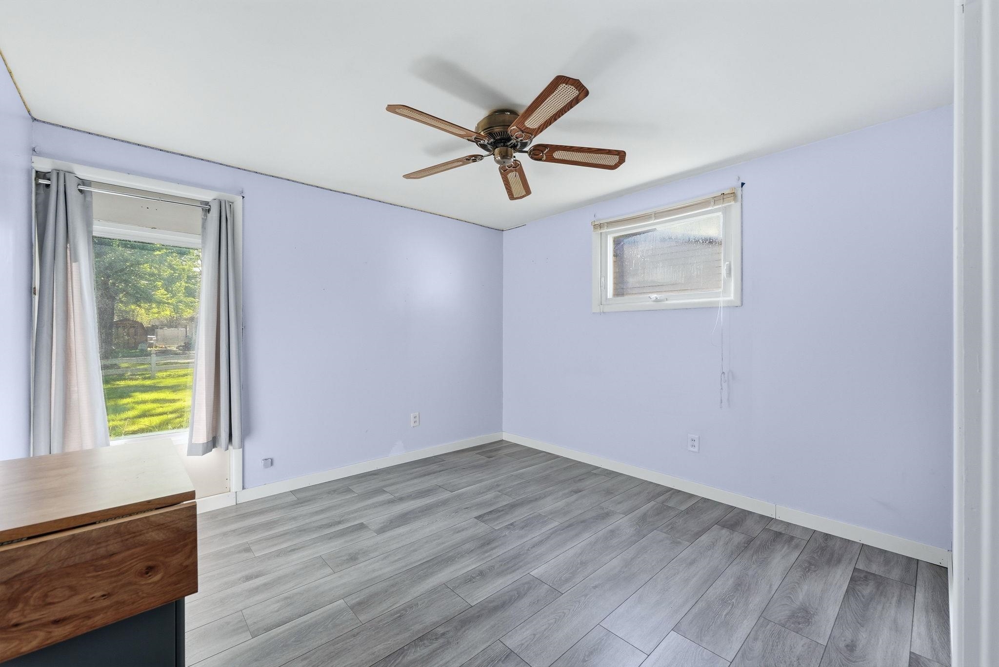 Spacious room featuring wood-finish flooring, a ceiling fan with integrated lighting, two windows for natural light, and light-toned wall finishes