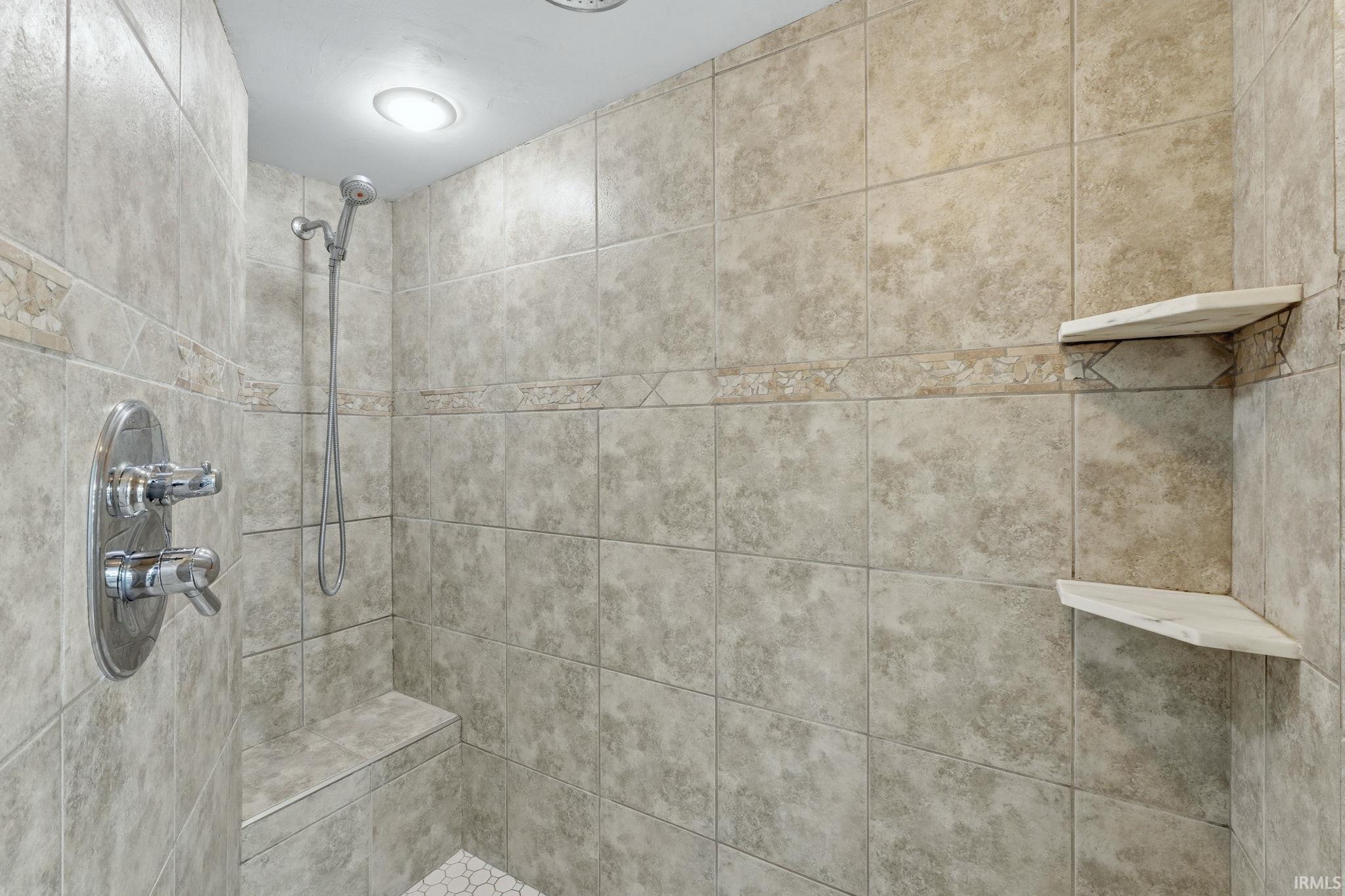 Tiled shower enclosure featuring a chrome shower fixture with handheld spray, integrated bench seating, recessed lighting, and dual corner storage shelves