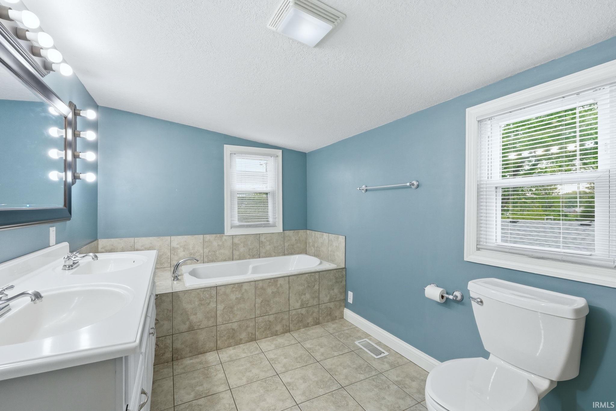 Bathroom featuring a double vanity with integrated sinks, a built-in bathtub with tiled surround, a toilet, a window with blinds, and tiled flooring