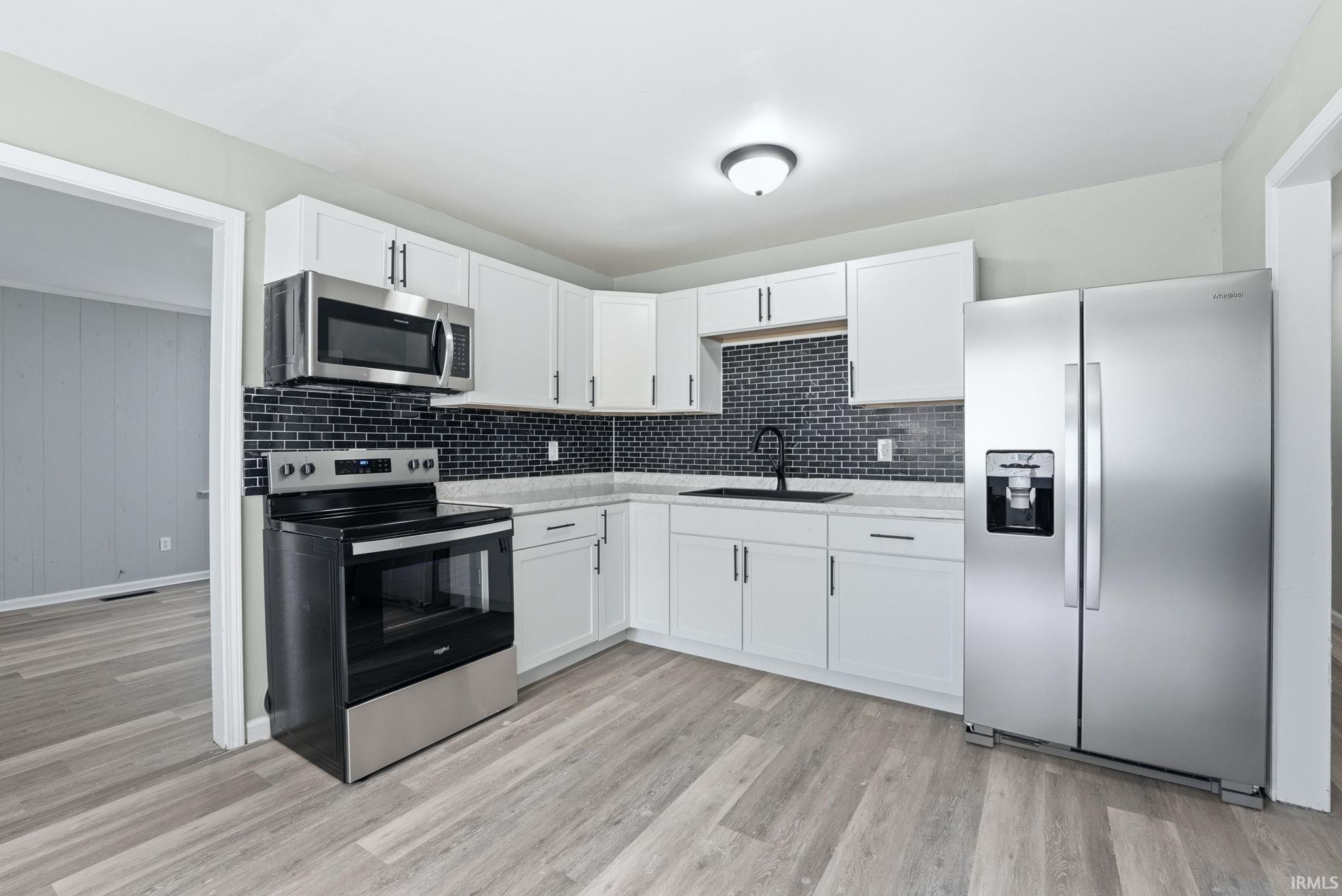 Modern kitchen featuring white cabinetry, black subway tile backsplash, stainless steel appliances, wood-finish flooring, and a contemporary ceiling light fixture