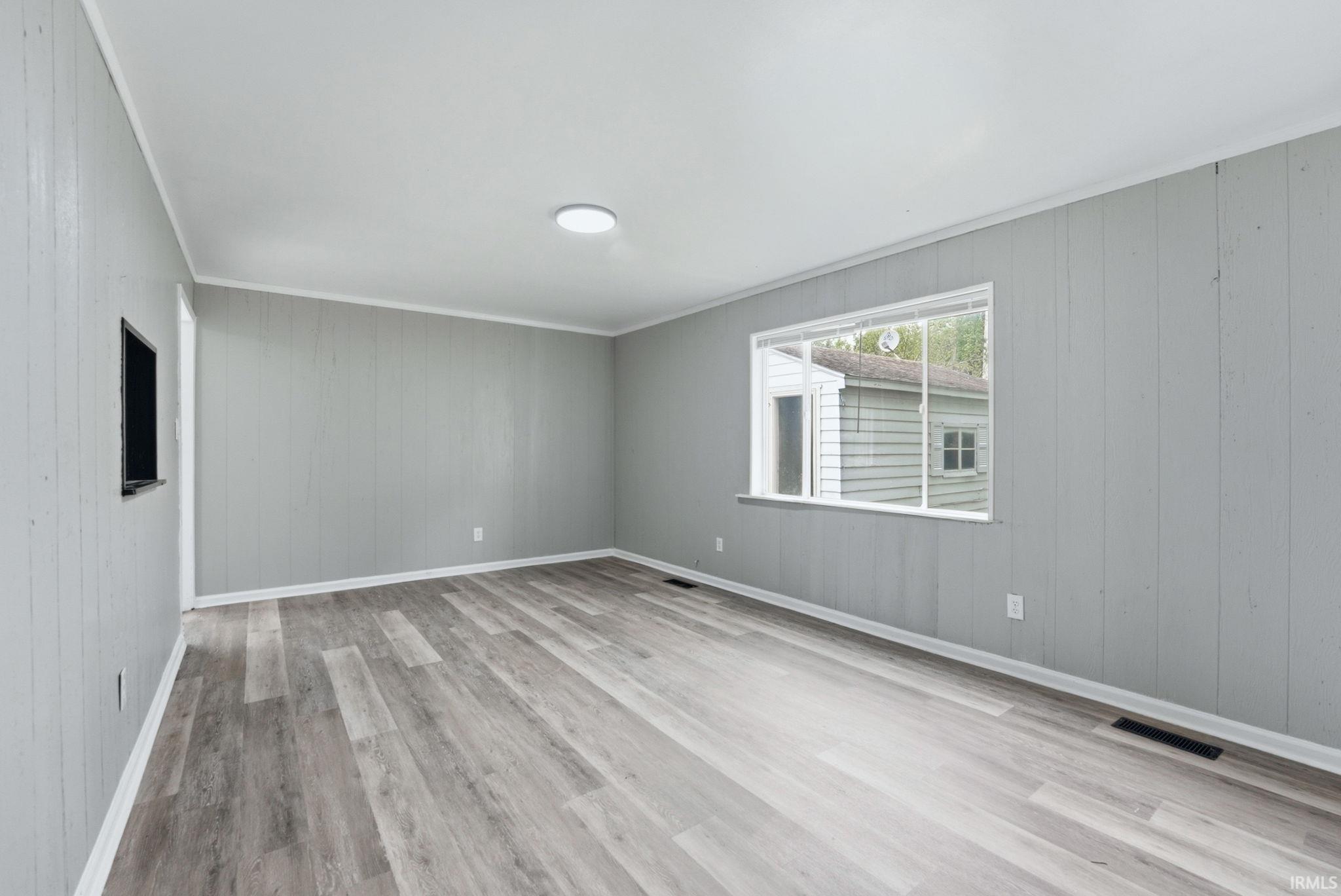 Spacious room featuring wood-finish flooring, light grey paneling, bright white trim, a large window, and a flush-mount ceiling light fixture