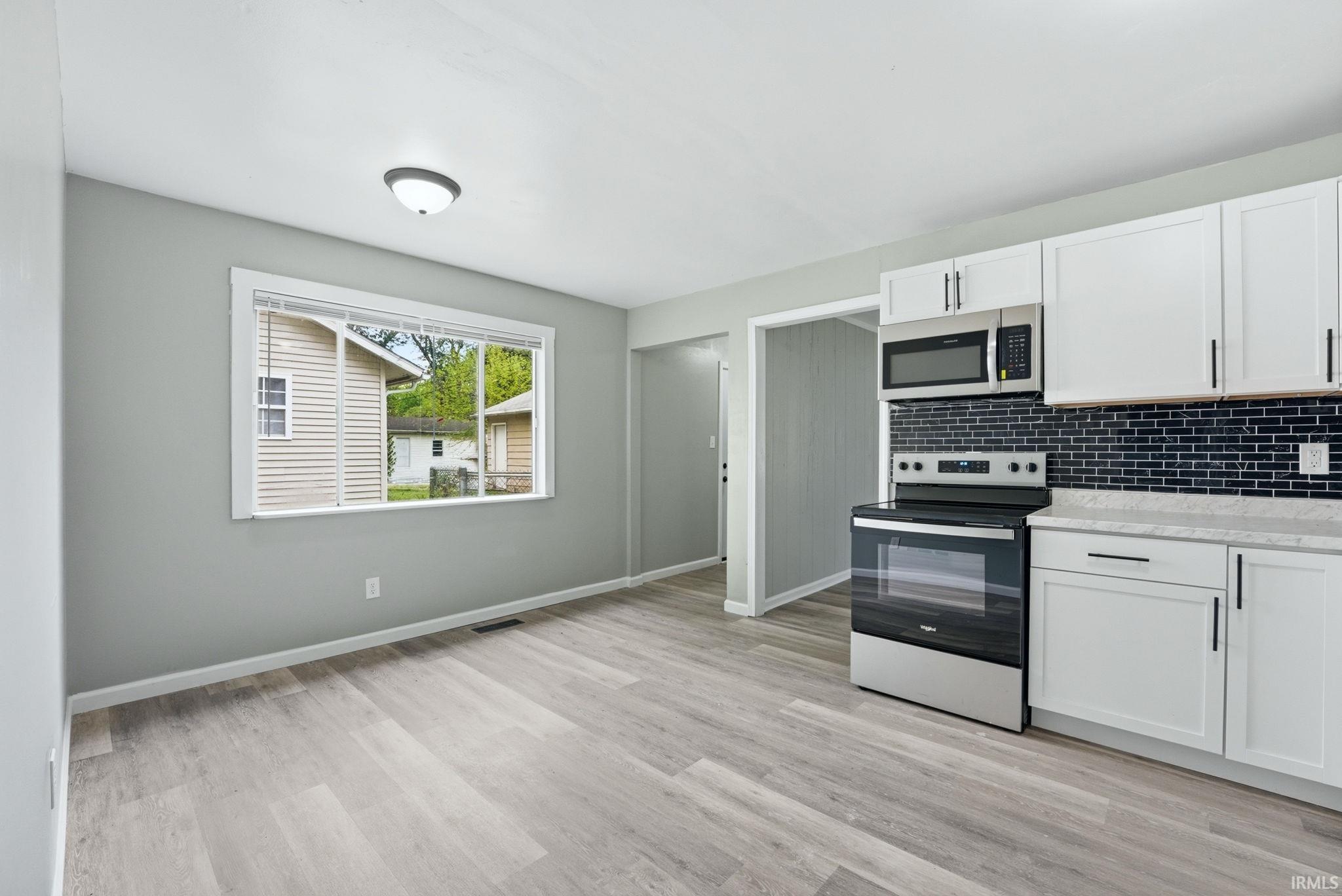 Kitchen space featuring wood-finish flooring, white cabinetry with black hardware, a stainless steel microwave and range, and a dark subway tile backsplash