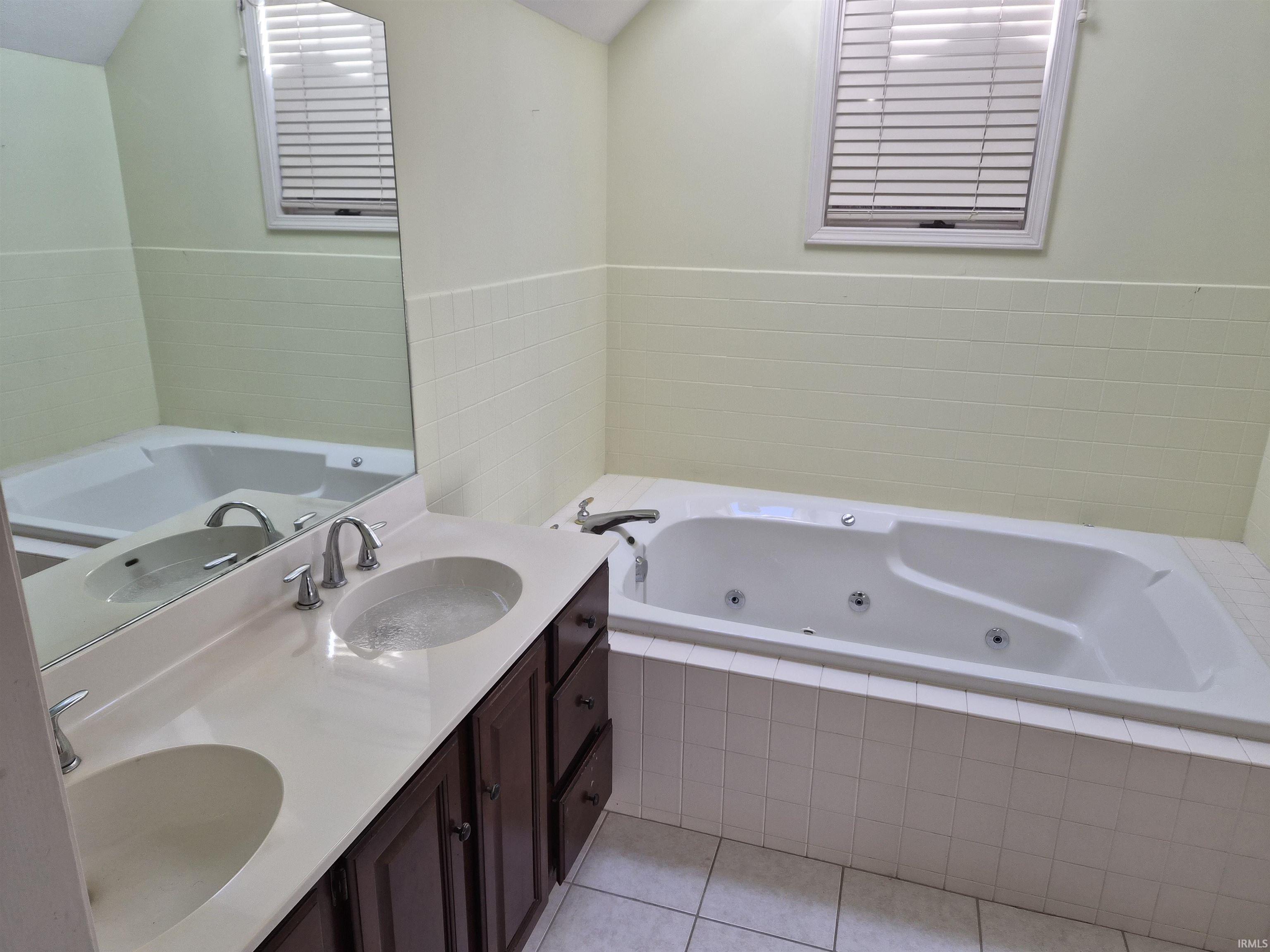 Bathroom featuring a built-in jetted tub, dual vanity with undermount sinks, wood-finish cabinetry, and tiled flooring