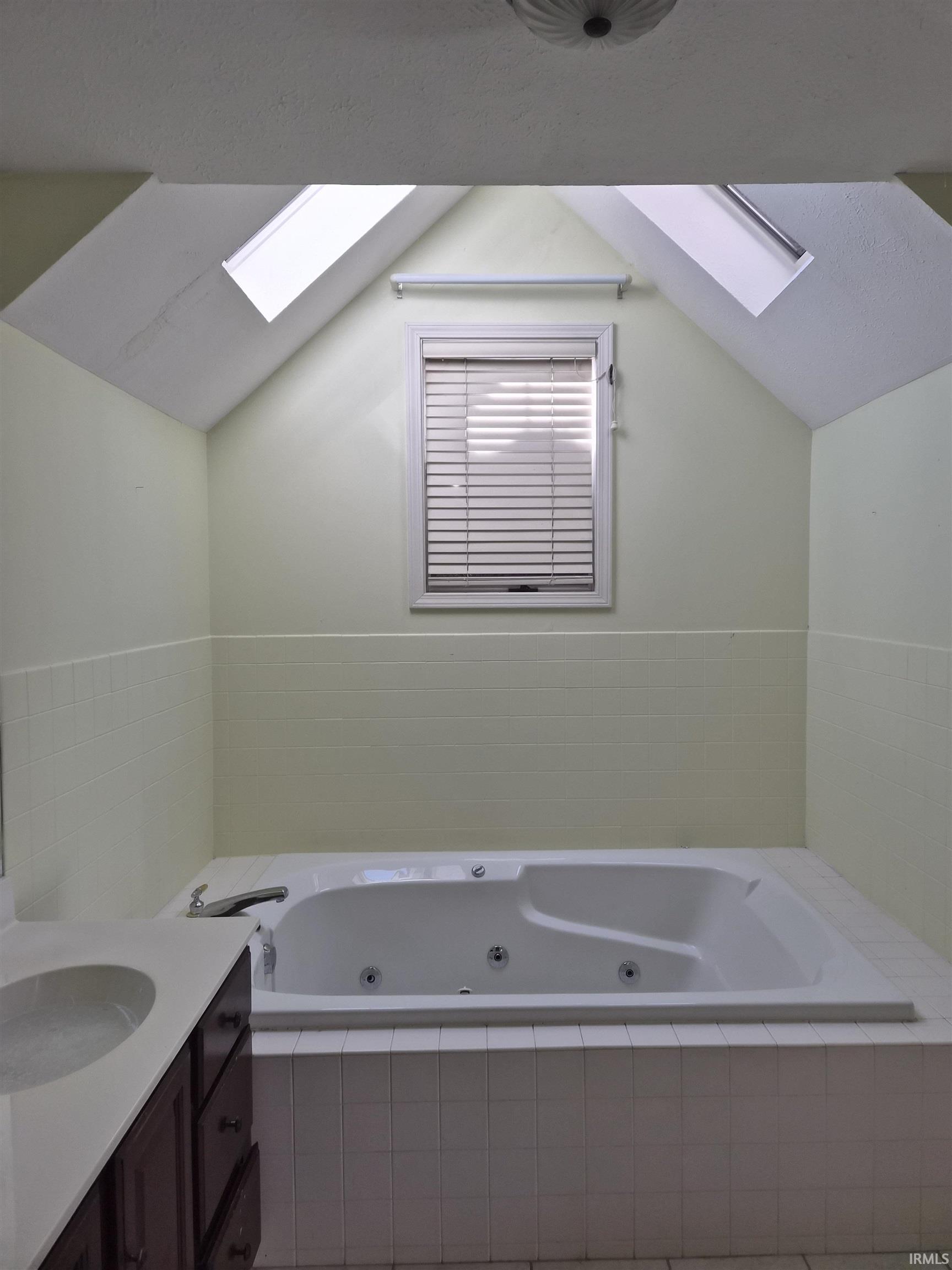 Soaking tub with integrated jets set within a tiled surround, featuring a window with blinds and two skylights