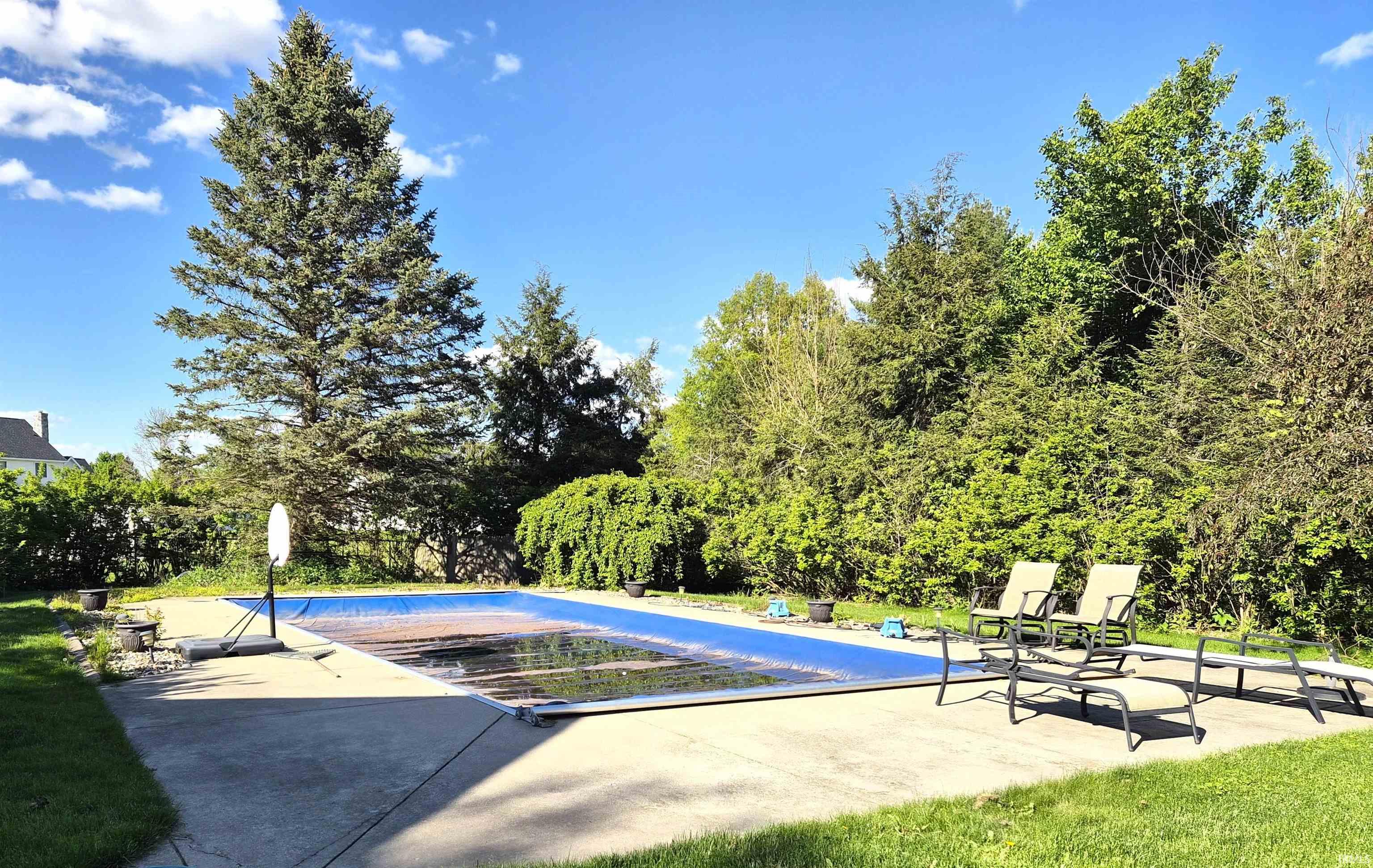 Expansive outdoor space featuring a rectangular swimming pool with a protective cover, a concrete pool deck, and surrounding mature trees and shrubs