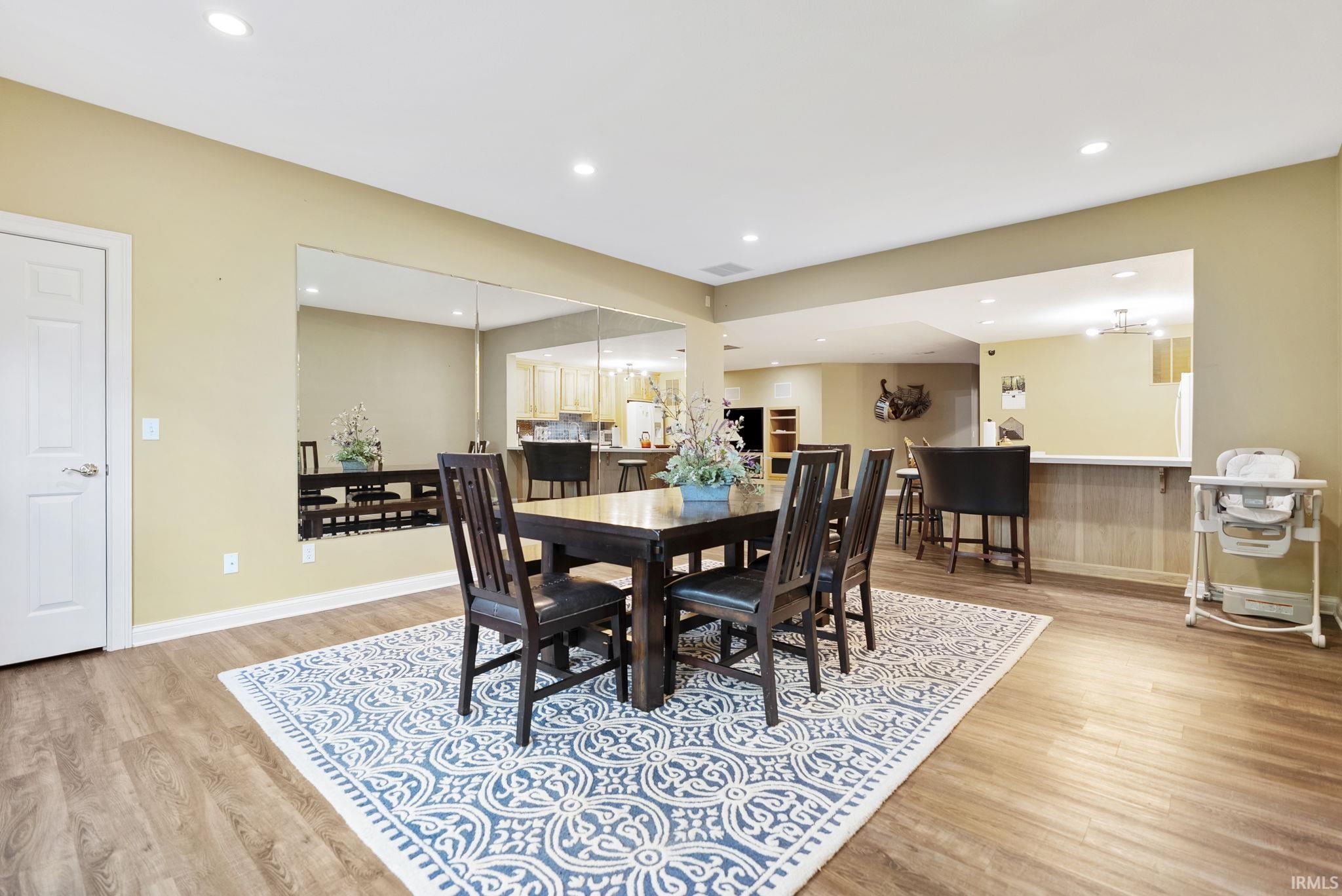 Spacious dining area featuring wood-finish flooring, recessed lighting, a full-height mirrored wall, and a pass-through opening to the kitchen