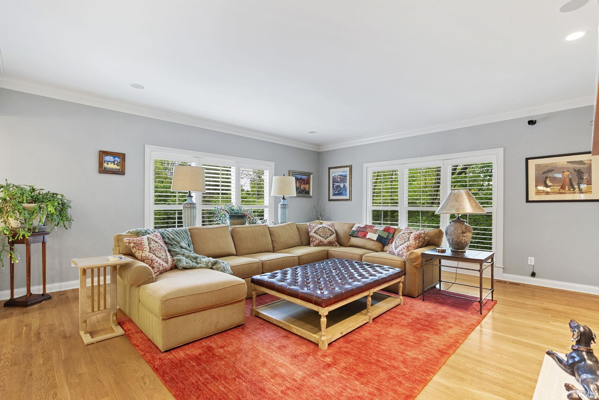 Spacious living area featuring wood-finish flooring, multiple windows with white plantation shutters, crown molding, and recessed ceiling lighting