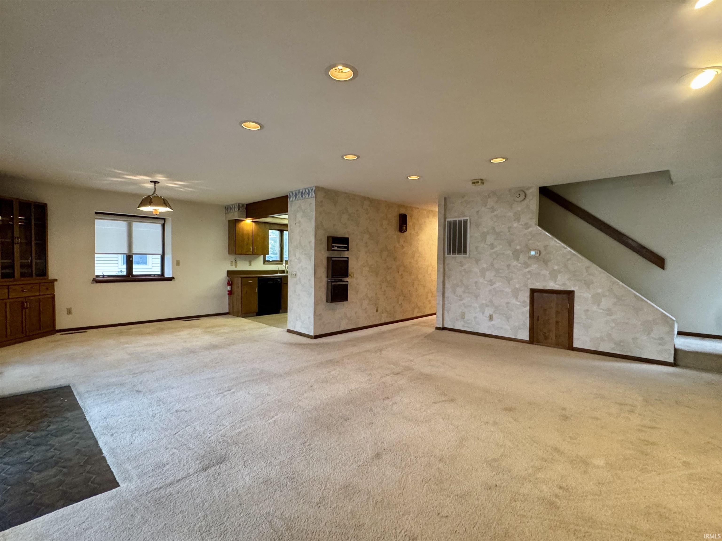 Expansive carpeted living area with recessed lighting, a built-in wooden hutch with glass-front cabinets, and a window with blinds