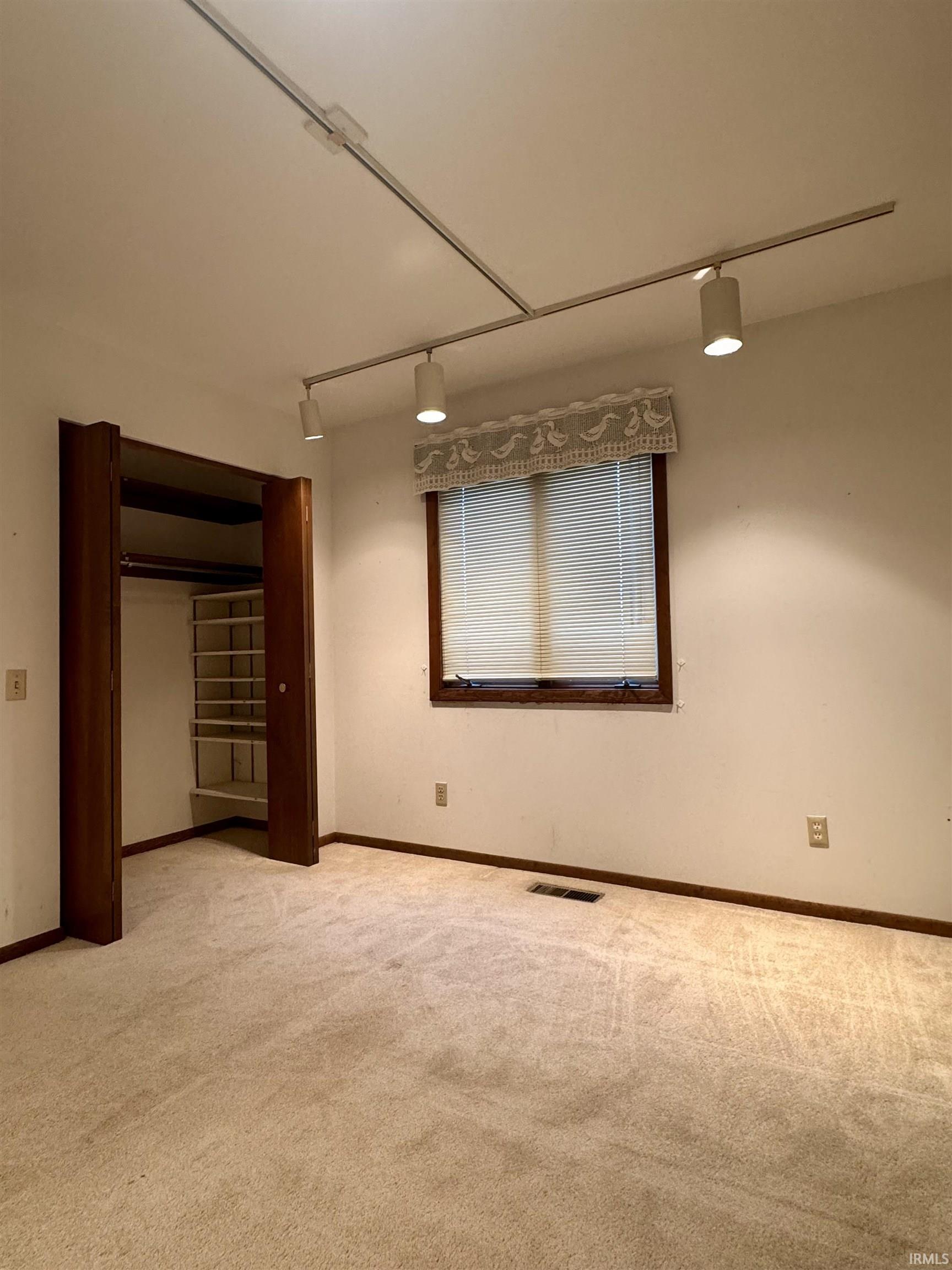 Carpeted room featuring a window with blinds, dark wood trim, track lighting, and a closet with built-in shelving