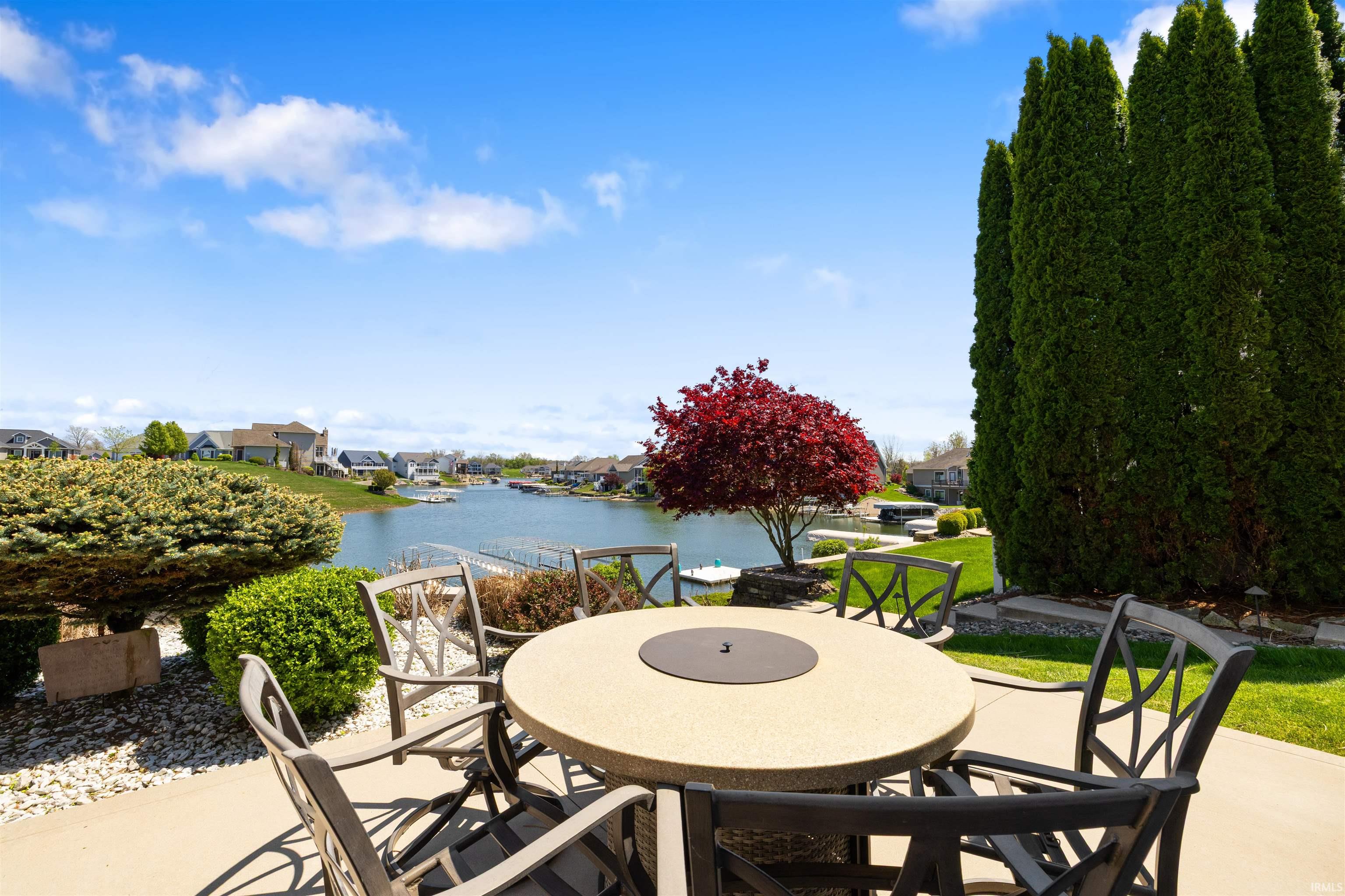 Concrete patio featuring a round composite fire pit table, manicured lawn, and mature evergreen shrubs