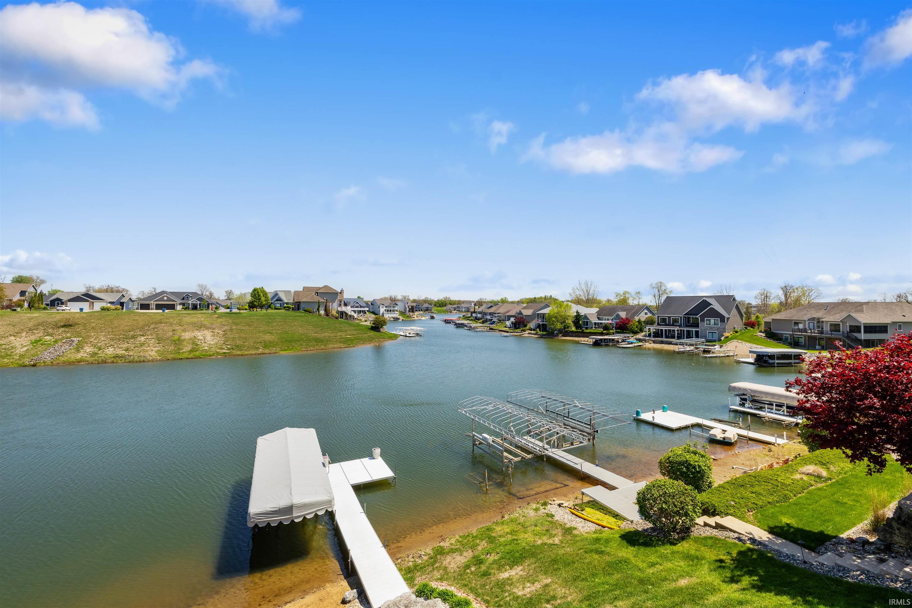 Waterside property featuring a private dock with covered boat lift, additional dock structure, sandy shoreline, manicured lawn, and mature landscaping