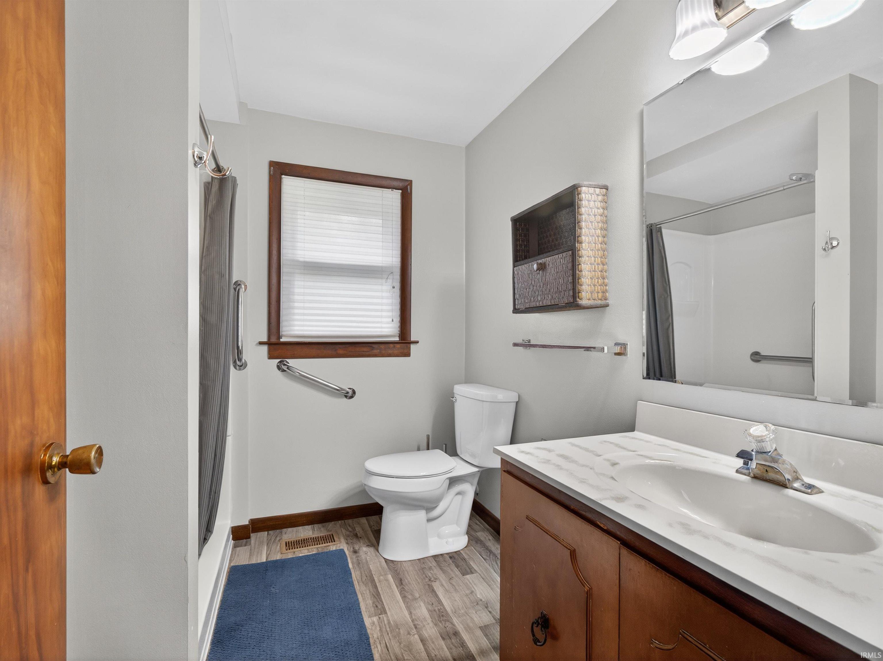 Bathroom featuring a wood-finish vanity with a single basin sink and a stone-look countertop, wood-finish flooring, a window with wood trim, and a shower with grab bars