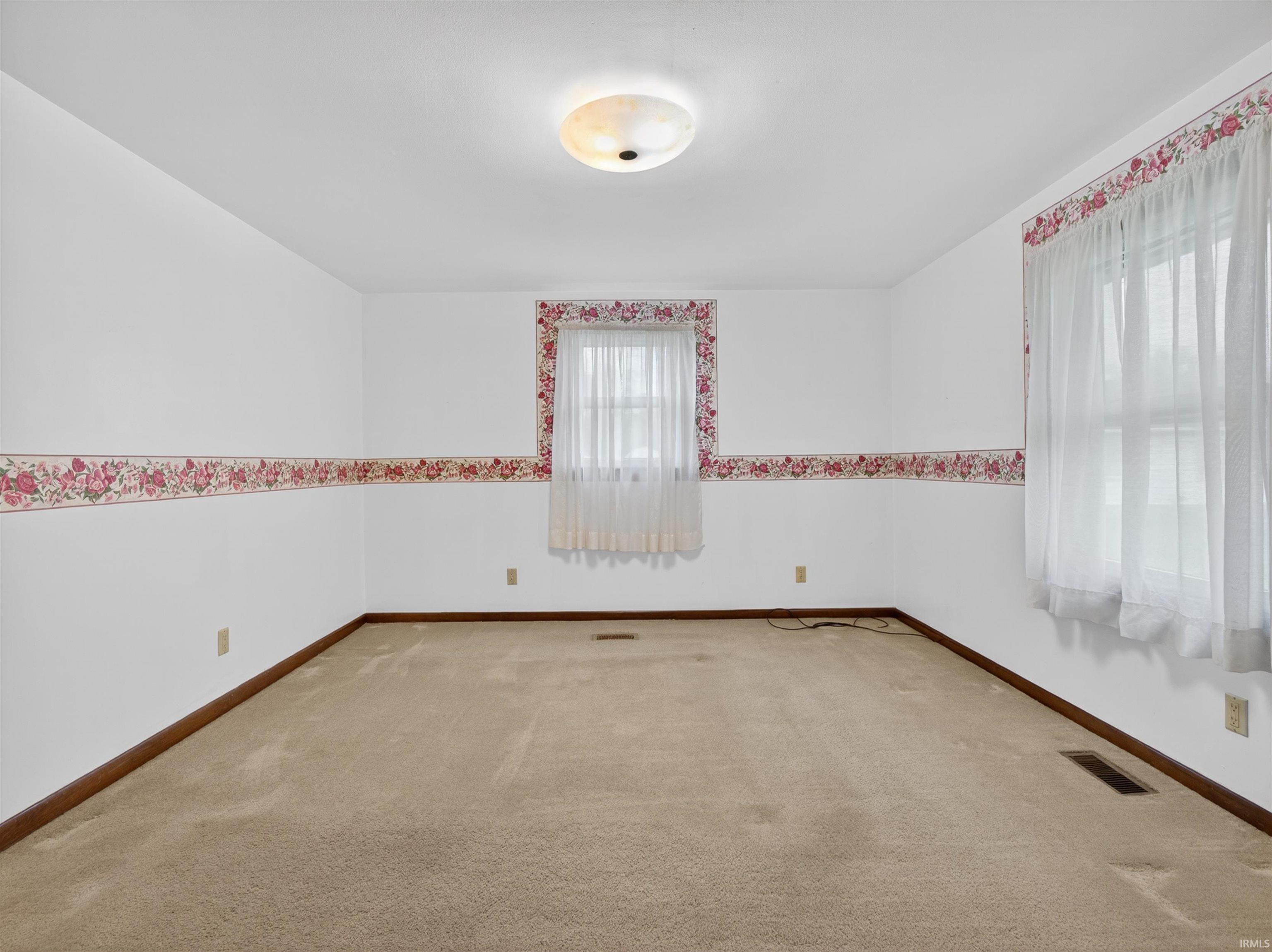Carpeted room featuring white walls with a floral border, two windows with white curtains, and a ceiling-mounted light fixture