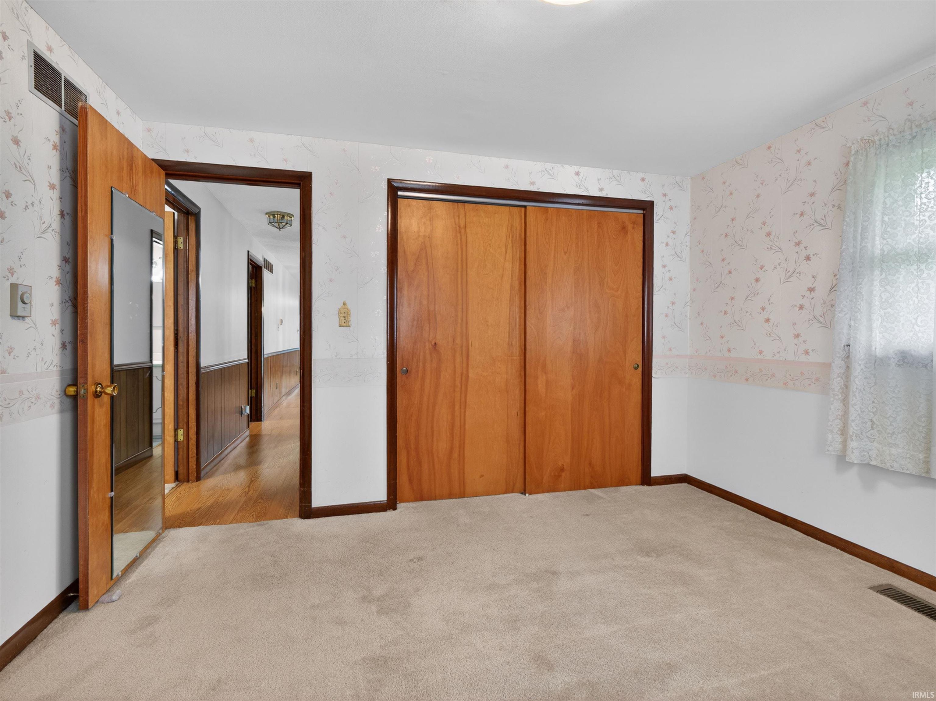 Carpeted room featuring wood-finish bi-fold closet doors, a window with sheer curtains, and wallpaper with a border
