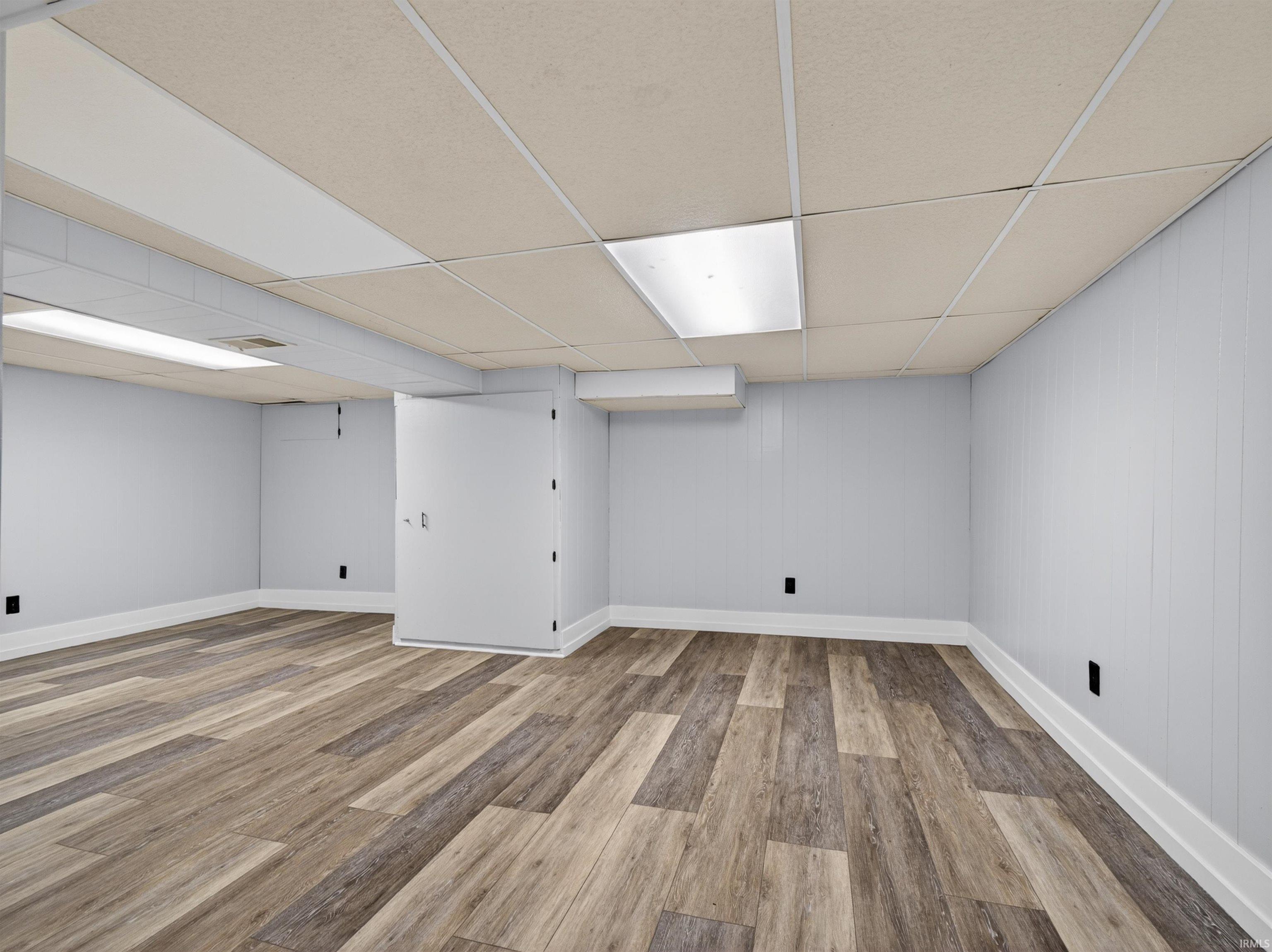 Spacious room featuring wood-finish plank flooring, light gray wall paneling, white baseboards, and a drop ceiling with integrated lighting