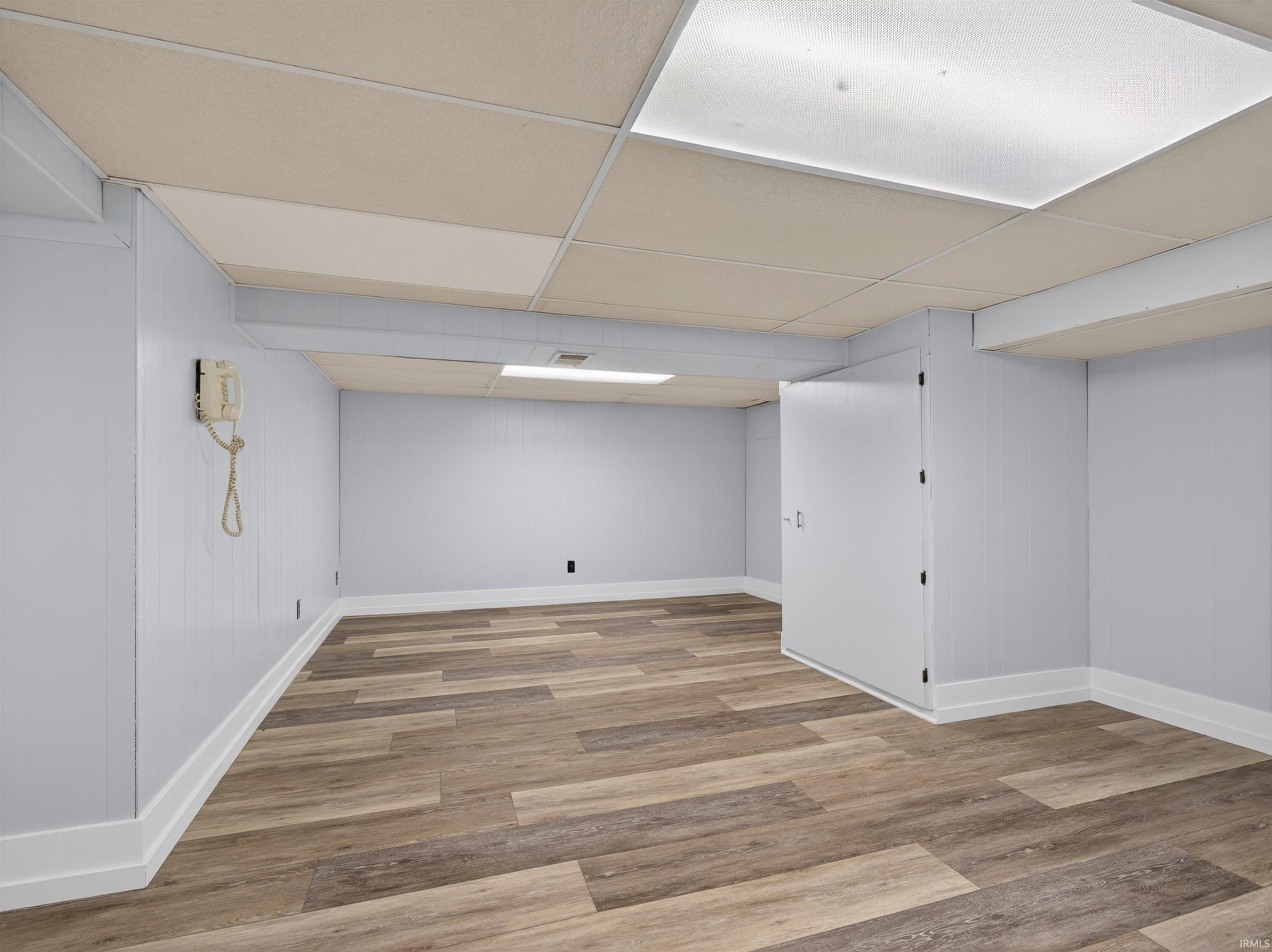 Spacious interior room featuring wood-finish flooring, light gray paneled walls, white baseboards, and a drop ceiling with integrated lighting