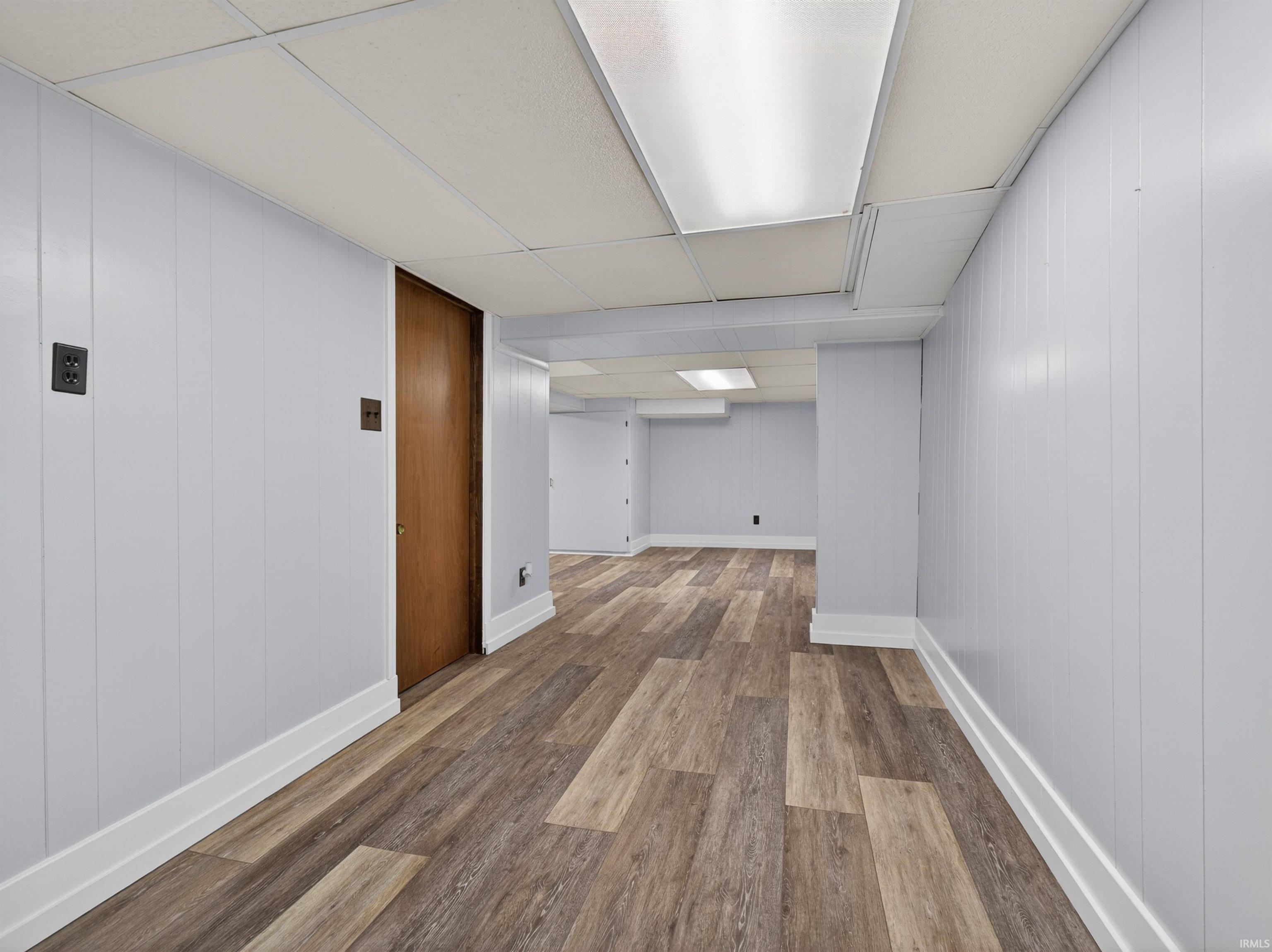 Spacious interior with wood-finish flooring, light-colored panel walls, white baseboards, and a dropped ceiling with integrated lighting