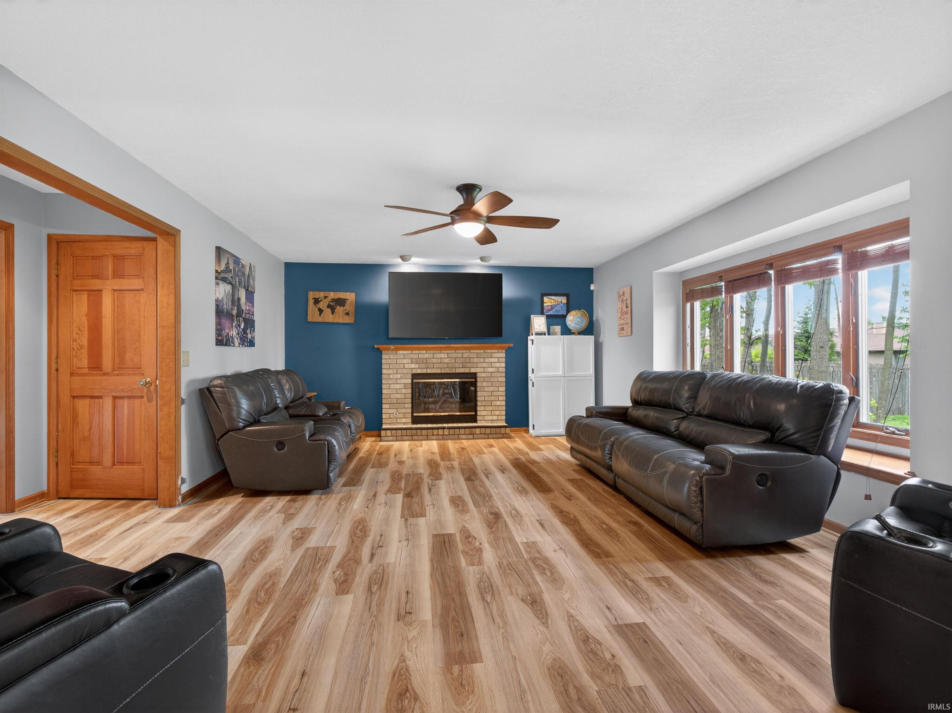 Spacious living area featuring wood-finish flooring, a brick fireplace with a wood mantle, and a ceiling fan with integrated lighting