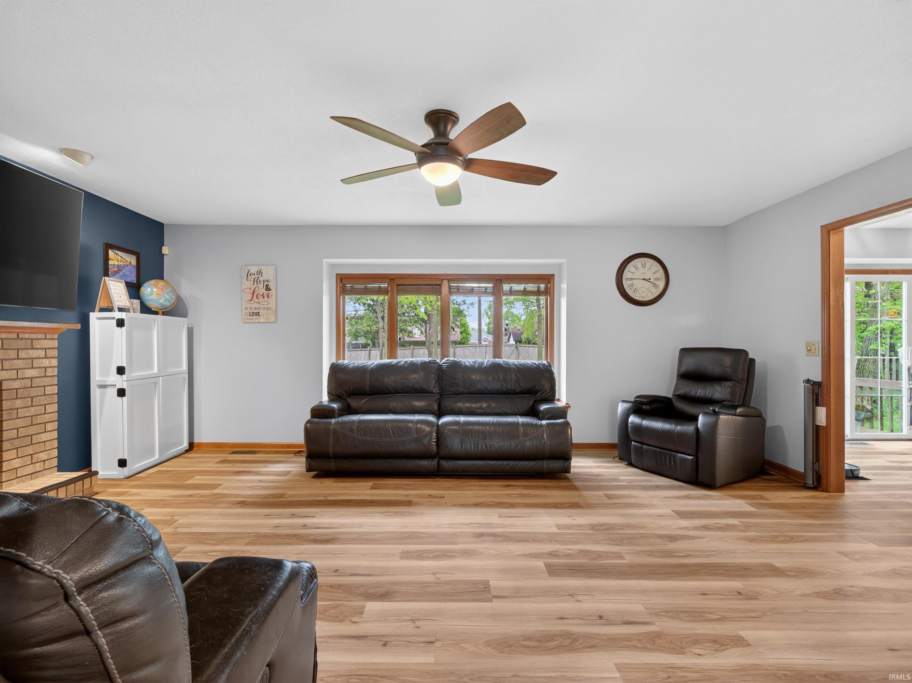 Spacious living area featuring wood-finish flooring, a ceiling fan with integrated lighting, and a brick fireplace