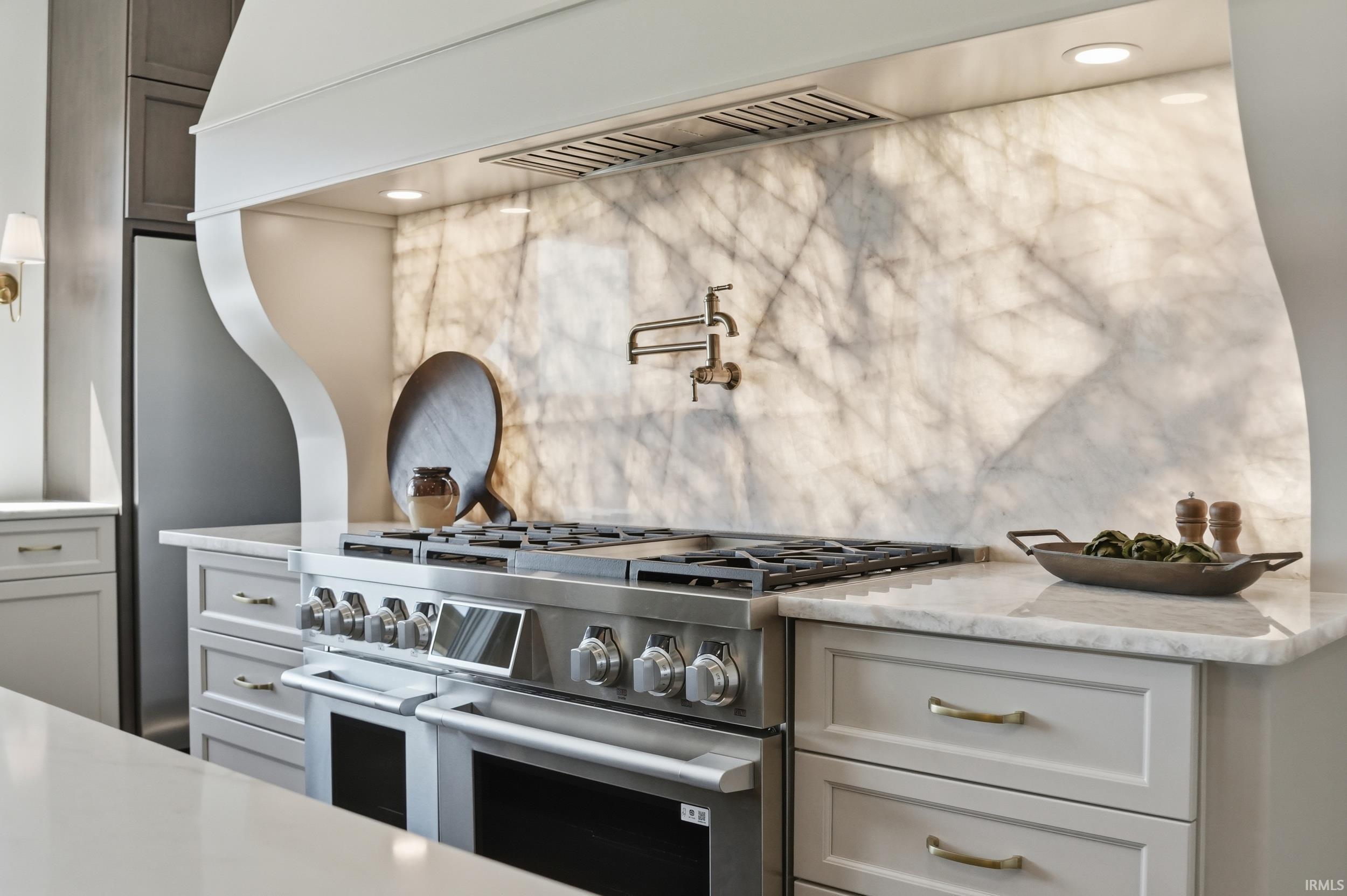 Gourmet kitchen featuring a professional-grade stainless steel range with double ovens, a striking translucent stone slab backsplash, custom range hood with integrated lighting, a brass pot filler, and light-toned cabinetry with brass hardware